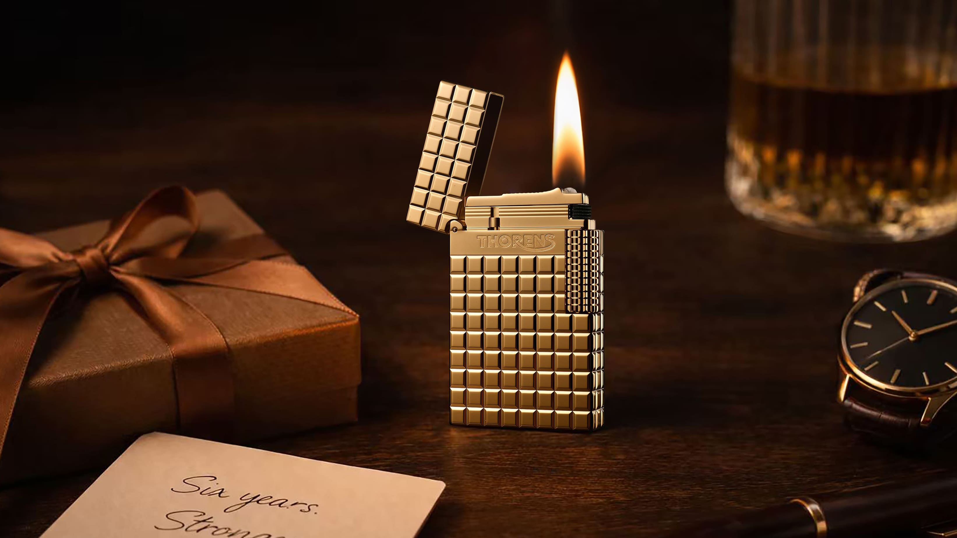 THORENS Side Slip Crisp Sound Auric Blocks fluid lighter displayed lit on a wooden table with a gift box, watch, and whiskey glass
