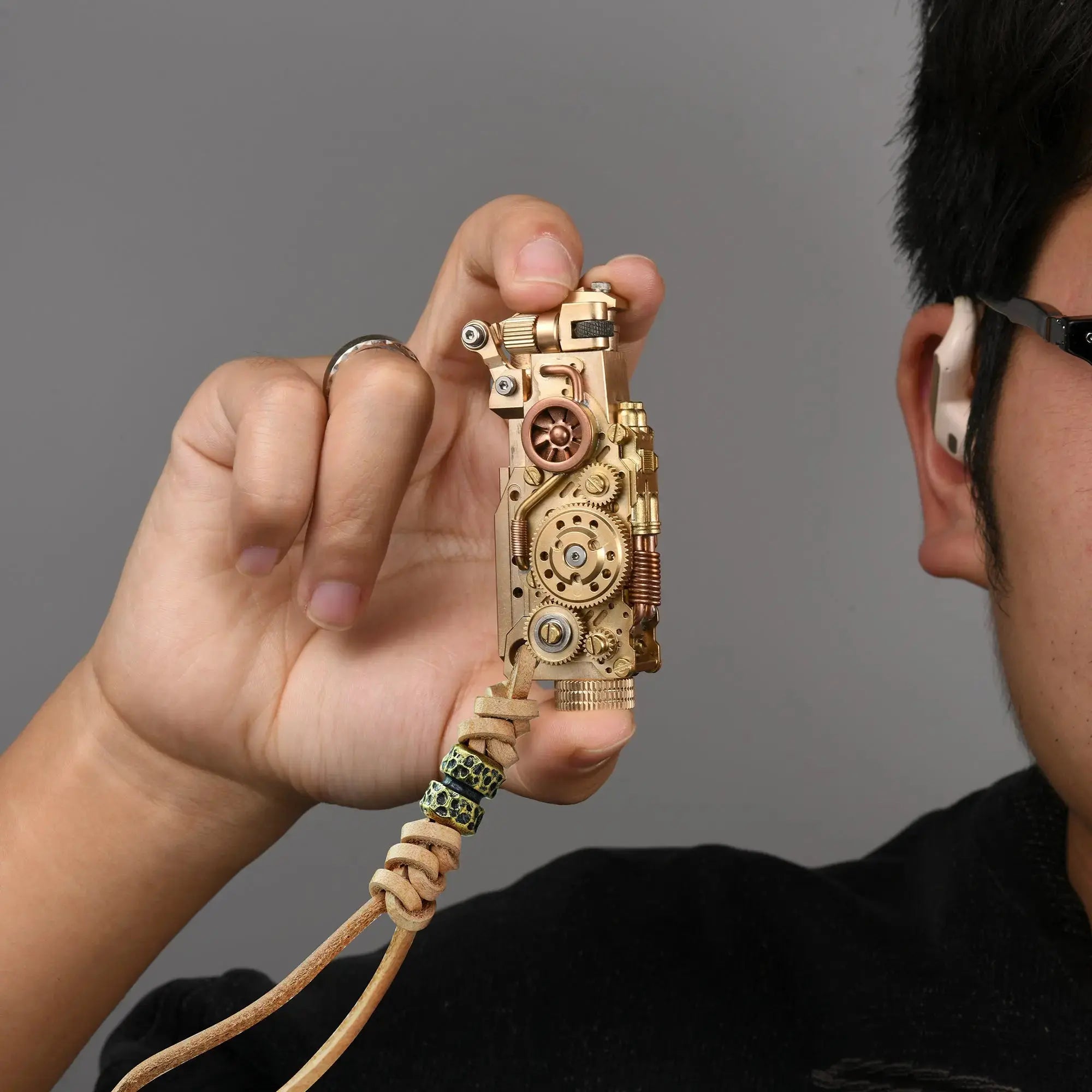 Thorens steampunk mechanical grip grand gear lighter in brass finish held in hand, featuring oversized exposed gear detailing, layered brass construction, and a leather lanyard with bead accents against a neutral background