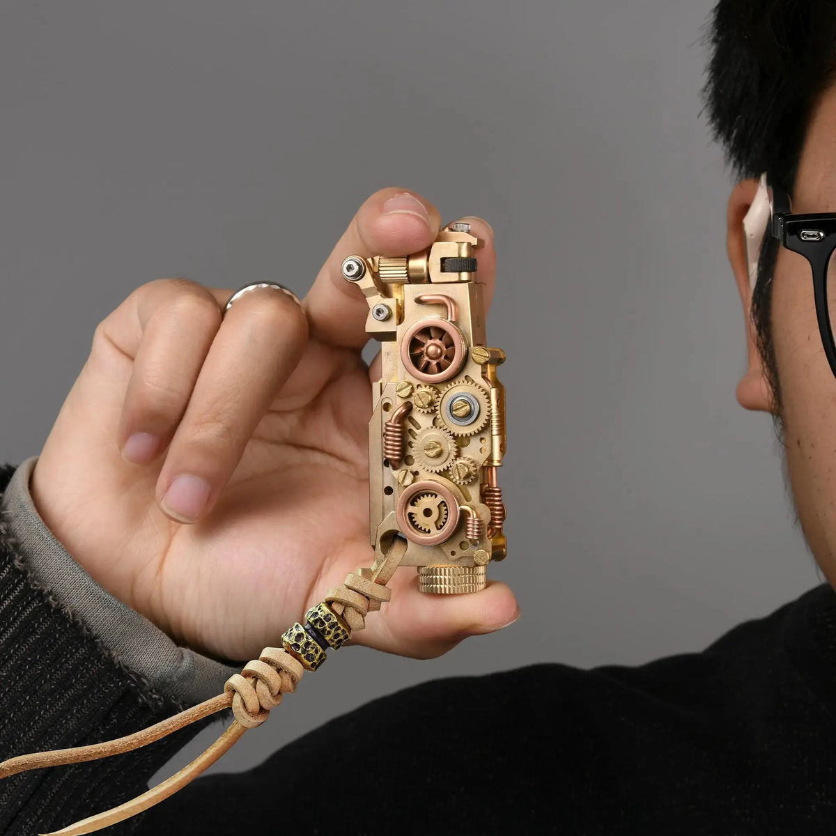 Thorens steampunk mechanical grip lighter in brass finish held in hand, featuring exposed fine gears, layered brass detailing, and a leather lanyard with bead accents against a neutral background