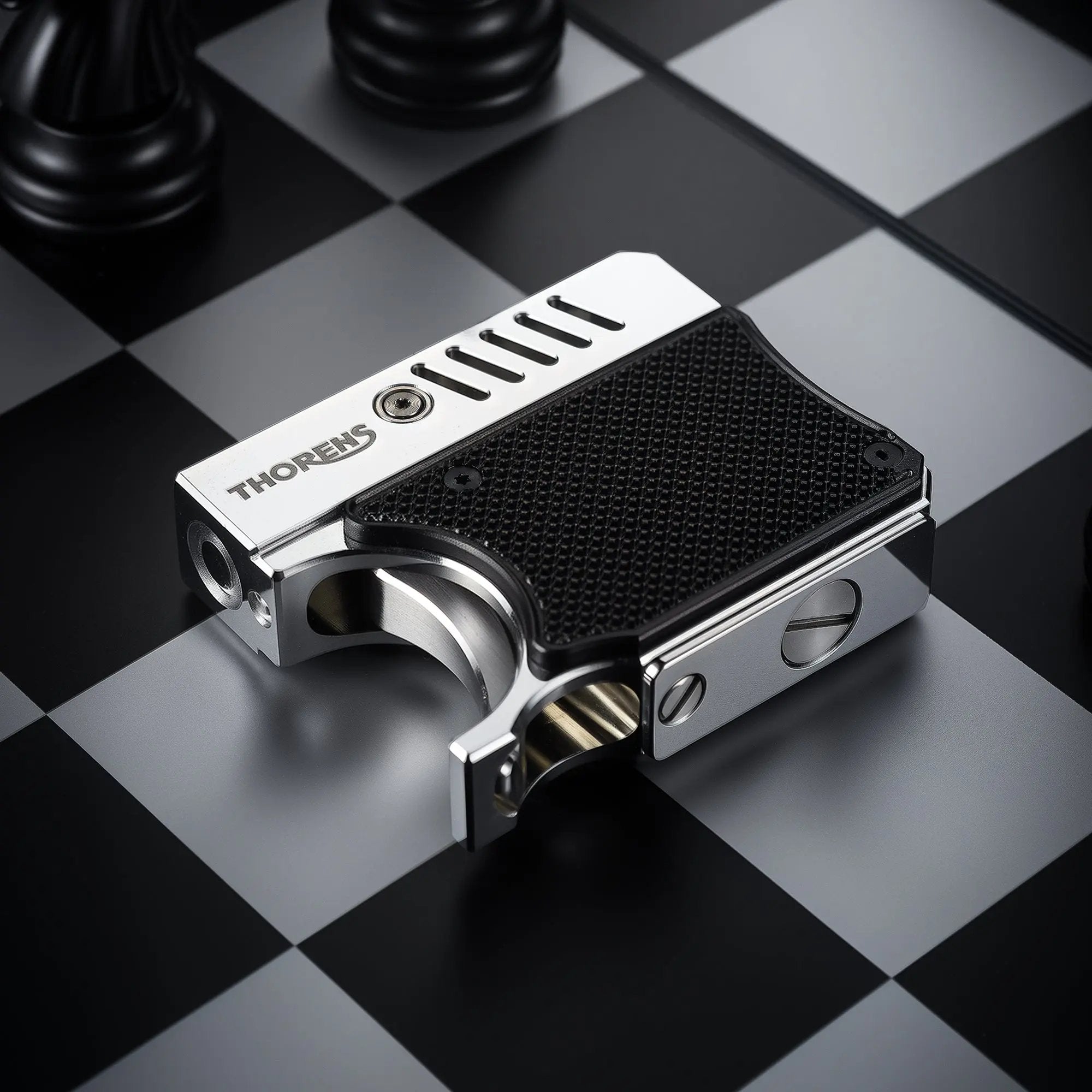 A side-angle view of the Thorens Gun Lighter on a chessboard, showcasing the metallic finish, the "THORENS" logo, vented cooling slots, and the textured black grip panel.