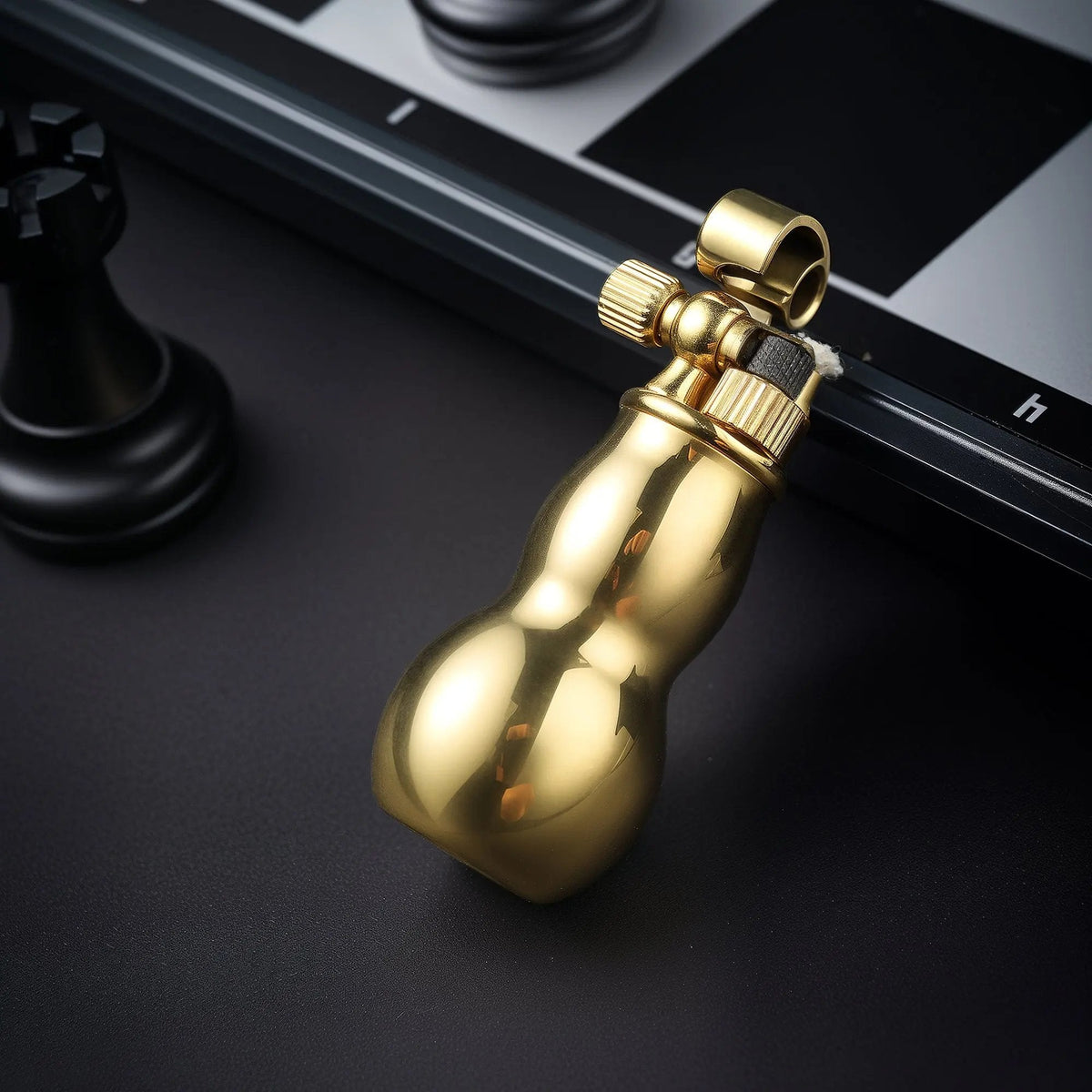 A polished brass Mini Gourd lighter resting on a dark surface next to a chessboard, showing the open mechanical cap and textured ignition wheel.
