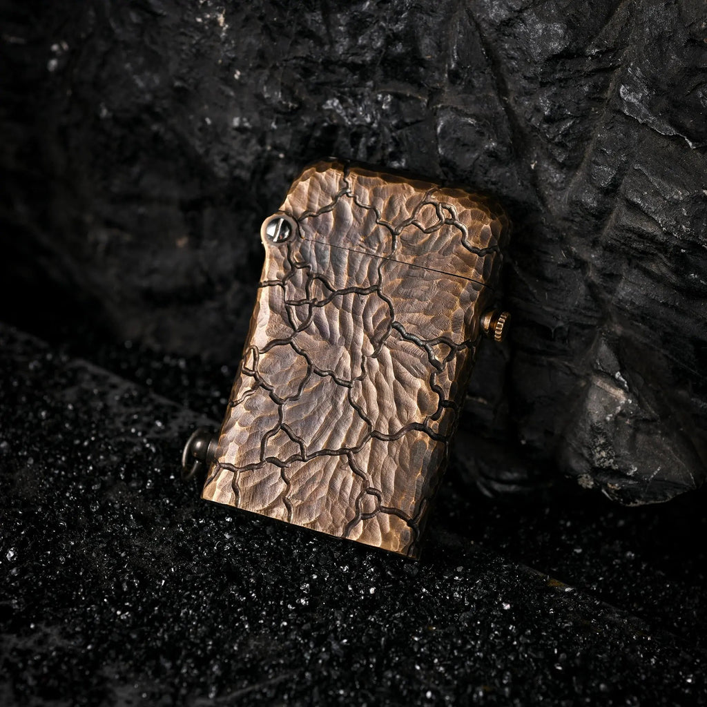 A Gen 1 Single Claw lighter with a hand-carved Earth Fissure texture, resting on black volcanic sand against a dark, craggy rock backdrop.