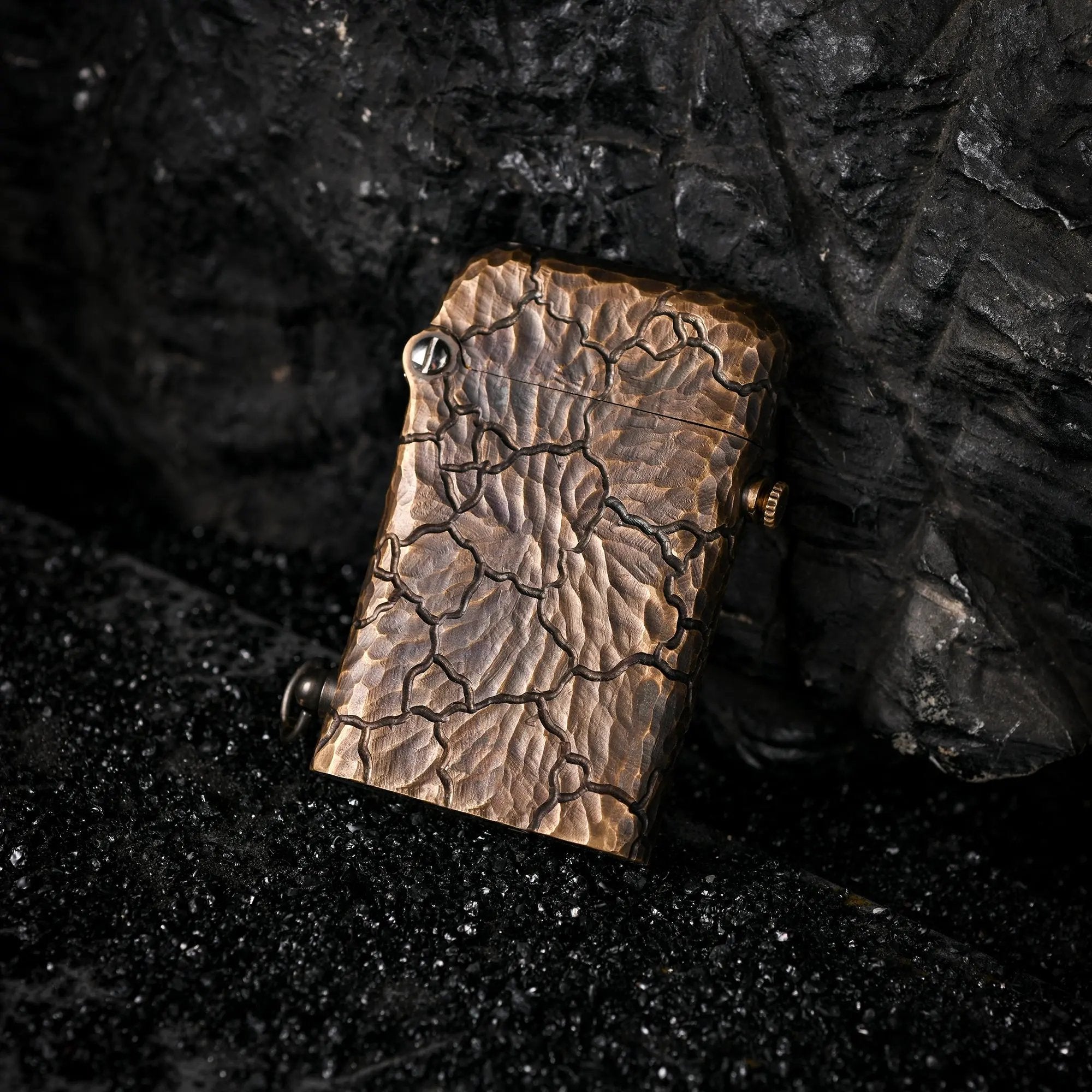 A Gen 1 Single Claw lighter with a hand-carved Earth Fissure texture, resting on black volcanic sand against a dark, craggy rock backdrop.
