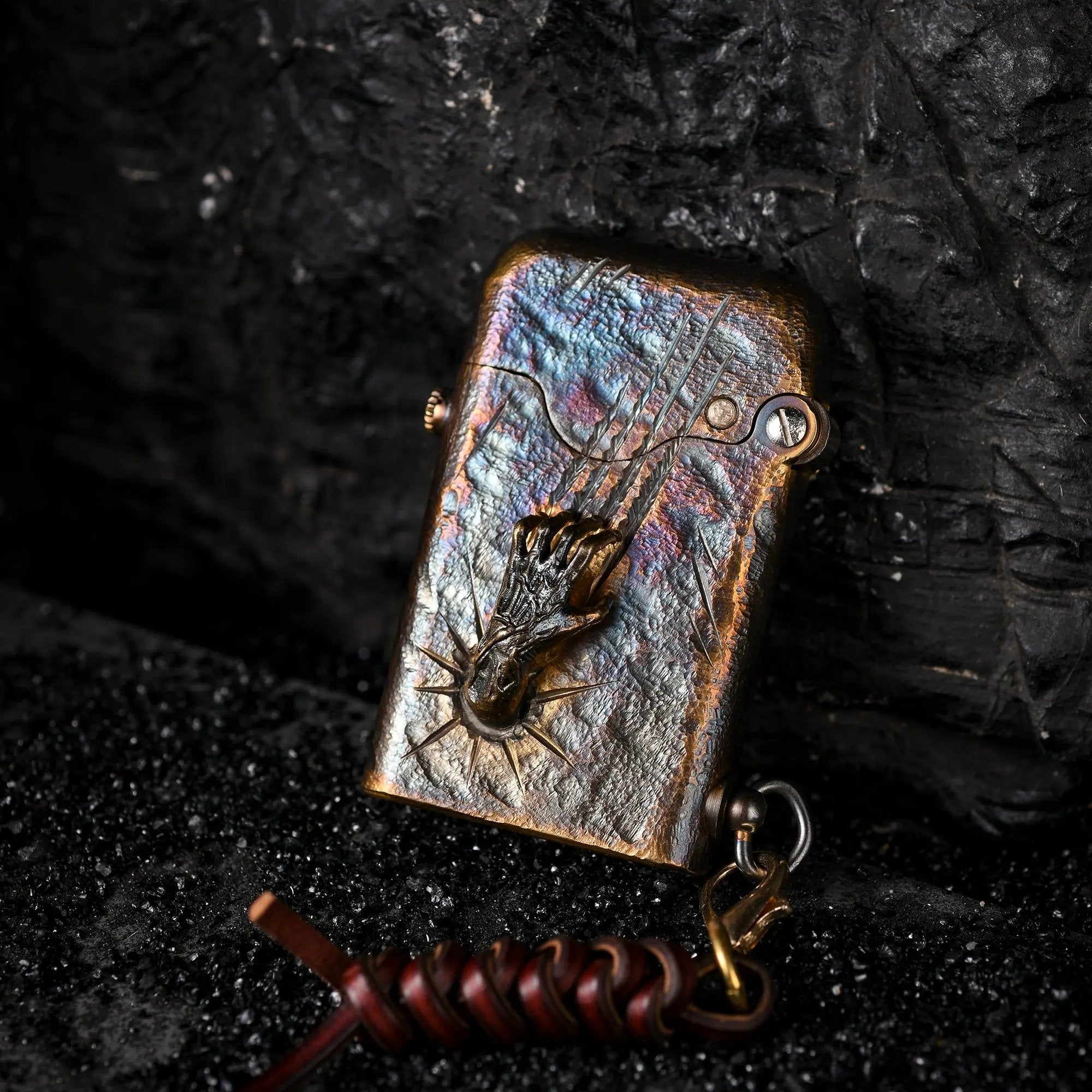 A Gen 1 Single Claw Spirit Claw lighter featuring a 3D skeletal hand relief and iridescent heat-treated brass, resting against dark volcanic rock.