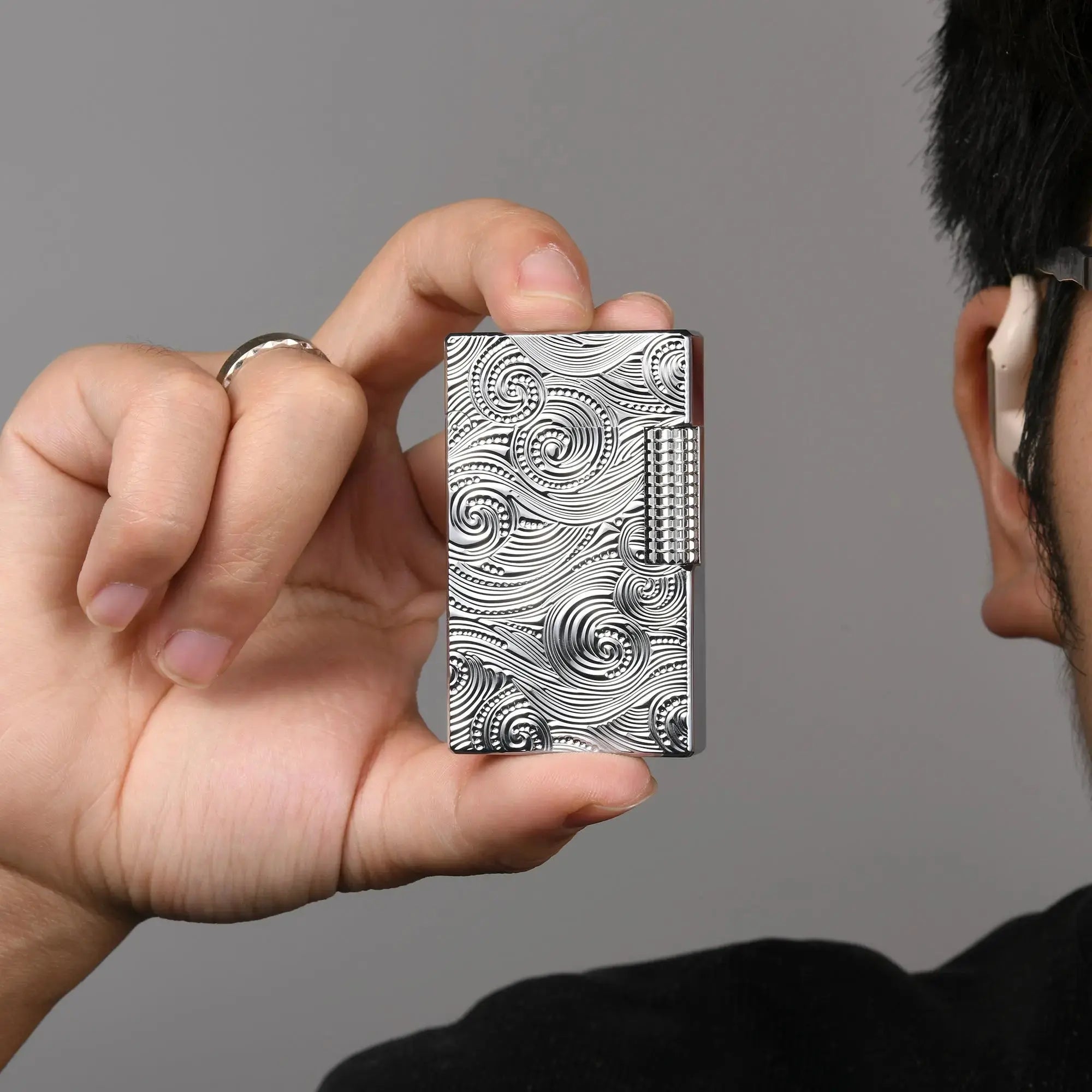 A person holding a silver Thorens side slip lighter between their fingers, showcasing its compact size and the intricate vortex engraved pattern on the brass casing.