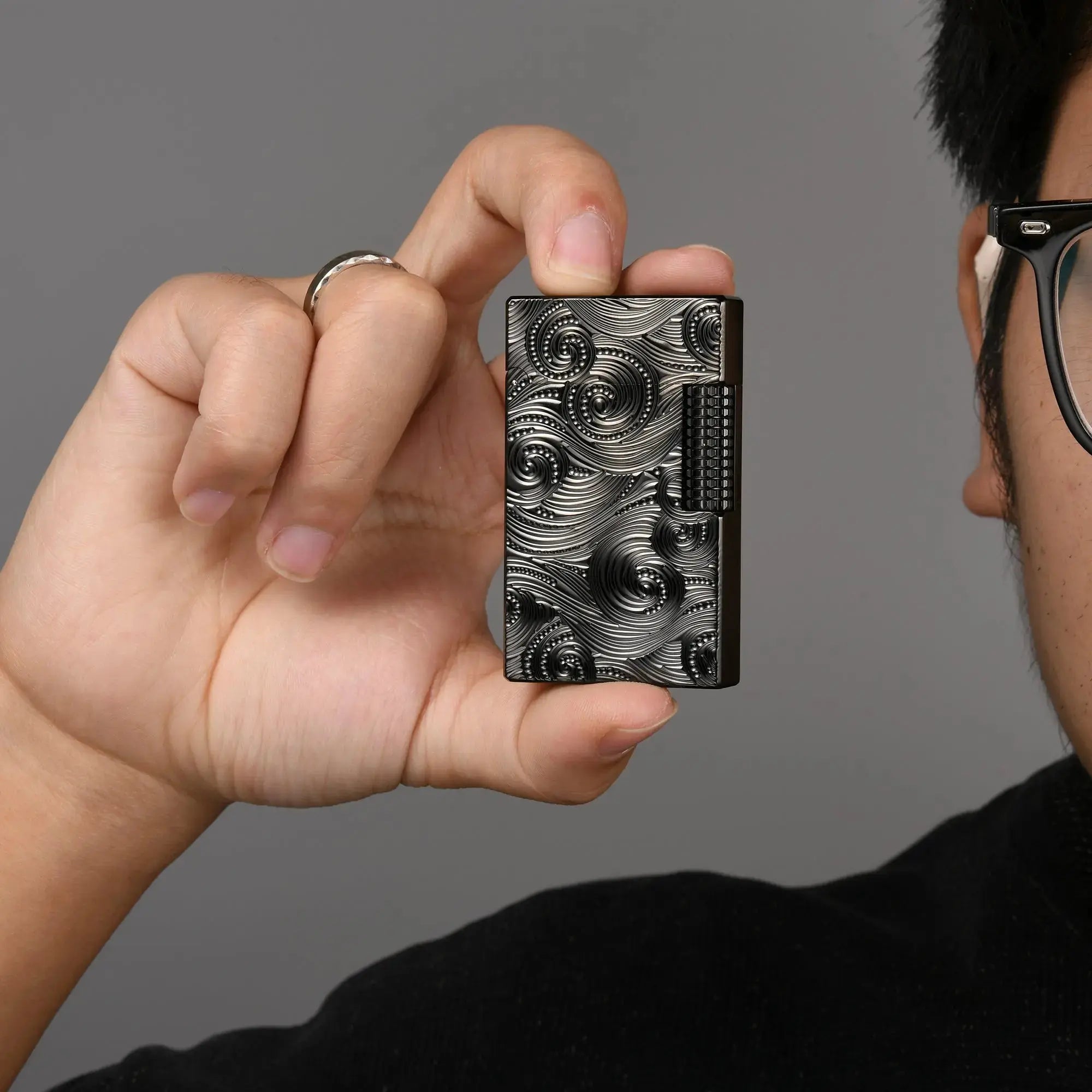 A person holding the Thorens Side Slip lighter in black Vortex engraved finish, demonstrating its compact size and refined EDC ergonomics. The light reflects off the deep-etched brass textures, showcasing the premium craftsmanship and mechanical side-slip flint wheel.