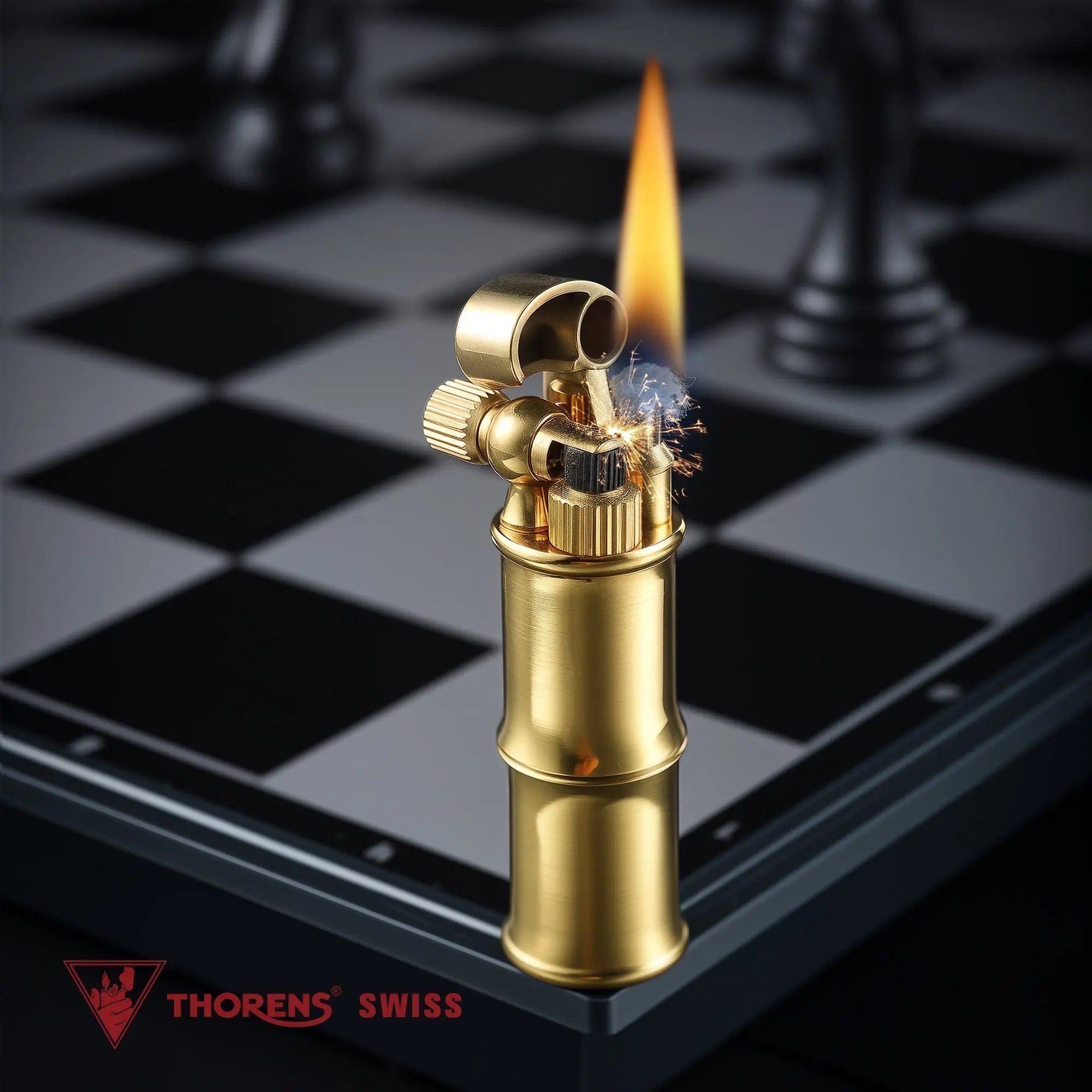 A brushed brass Mini Bamboo lighter producing a steady flame and visible ignition sparks, positioned on the edge of a silver and black chessboard with the Thorens Swiss logo in the foreground.