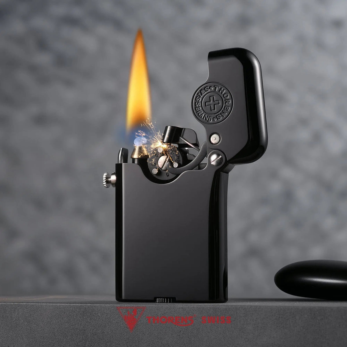 A lit Thorens Gen 3 single-claw lighter in glossy black, showing sparks and a bright flame against a soft-focus gray background with the Thorens Swiss logo.
