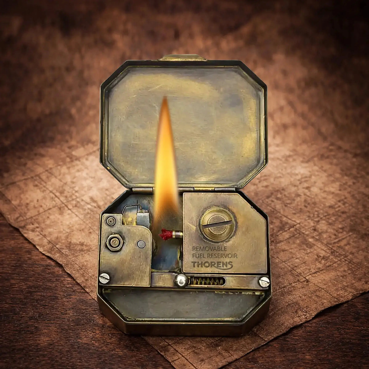 Thorens Knight’s Era matchbox lighter shown open with a visible flame, revealing the internal brass mechanism and removable fuel reservoir on a warm textured surface