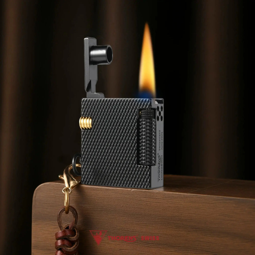 Thorens Mini Cube Black Mesh lighter shown lit on a wood surface, featuring a compact square form, textured black mesh finish, side flint wheel, gold-tone accent, and leather lanyard against a warm dark background