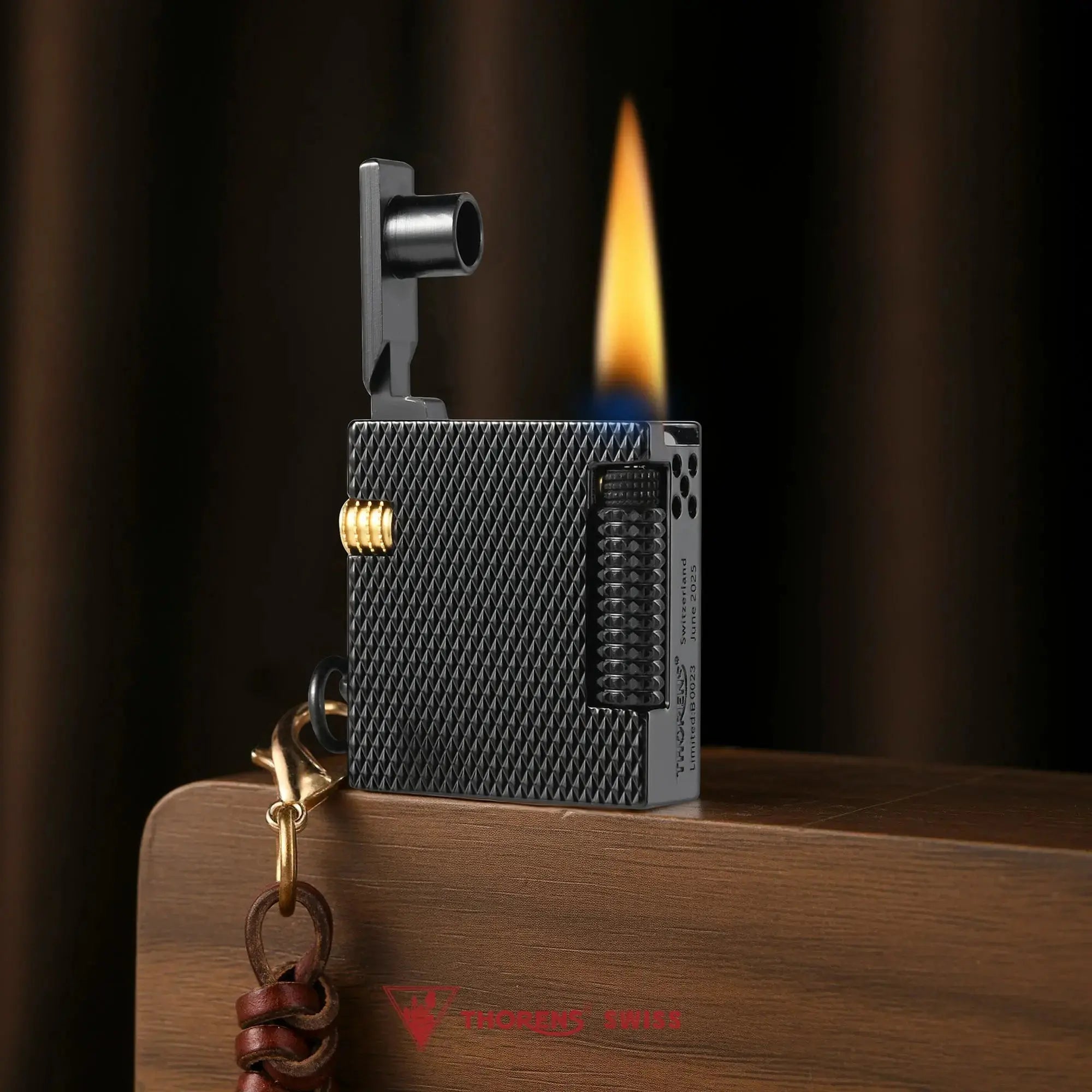 Thorens Mini Cube Black Mesh lighter shown lit on a wood surface, featuring a compact square form, textured black mesh finish, side flint wheel, gold-tone accent, and leather lanyard against a warm dark background