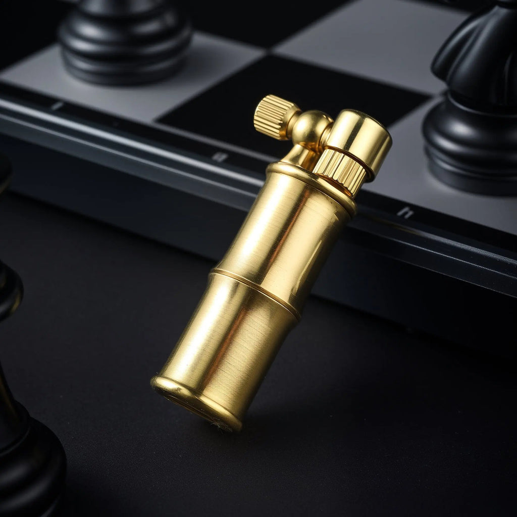 A compact brass lighter designed with a bamboo-stalk aesthetic and a side-mounted flint wheel, leaning against a black and silver chessboard with a chess piece in the background.