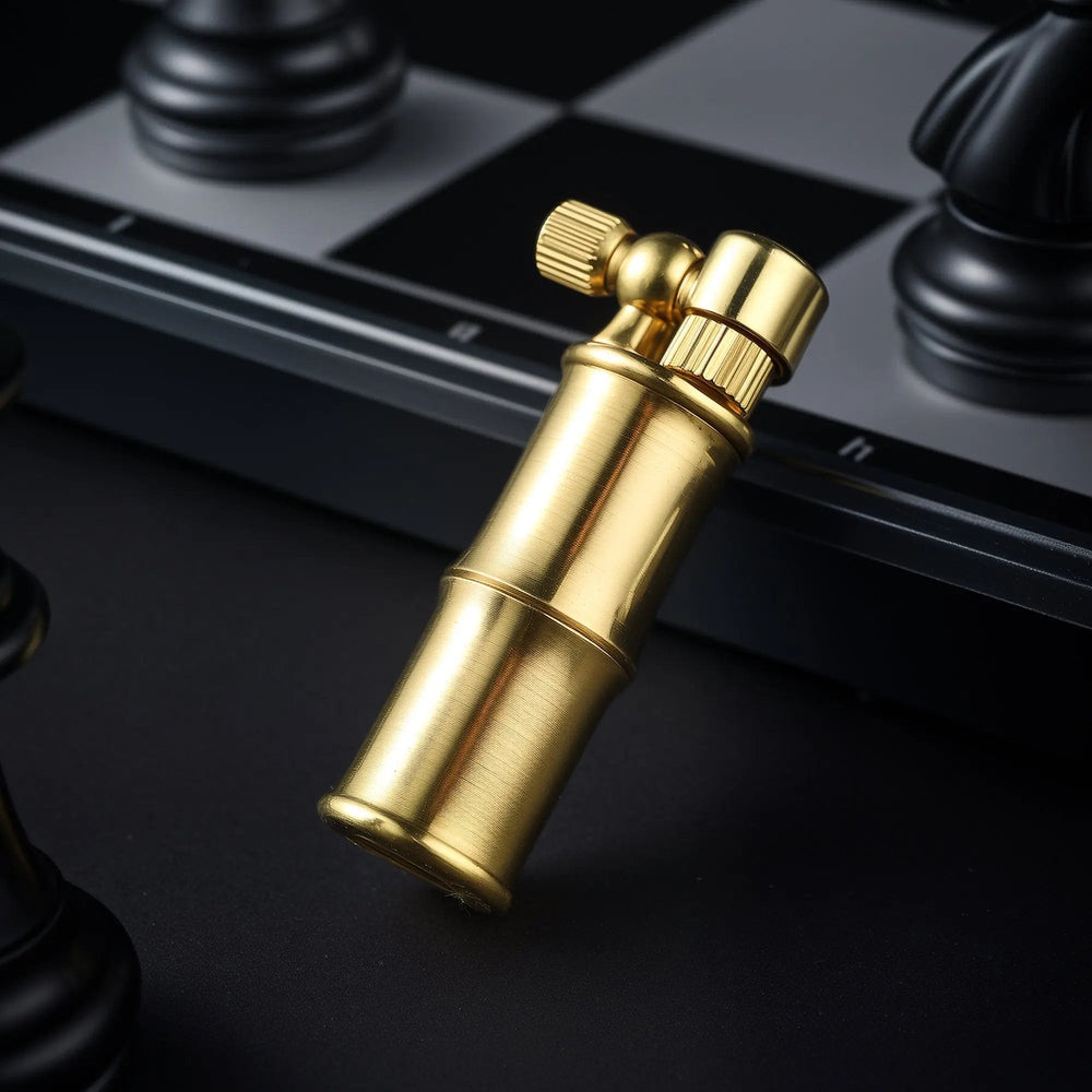 A compact brass lighter designed with a bamboo-stalk aesthetic and a side-mounted flint wheel, leaning against a black and silver chessboard with a chess piece in the background.