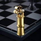 A vertical close-up of the Mini Bamboo brass lighter on a silver and black chessboard, showing the snuffer cap flipped open to reveal the cotton wick and mechanical ignition assembly.