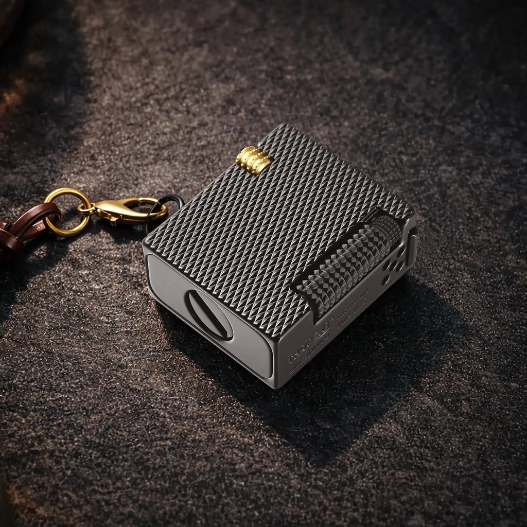 Thorens Mini Cube Black Mesh lighter resting on a dark textured surface, featuring a compact square form, black mesh pattern, silver-tone body, gold-tone accent, and attached leather lanyard hardware