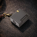 Thorens Mini Cube Black Mesh lighter resting on a dark textured surface, featuring a compact square form, black mesh pattern, silver-tone body, gold-tone accent, and attached leather lanyard hardware