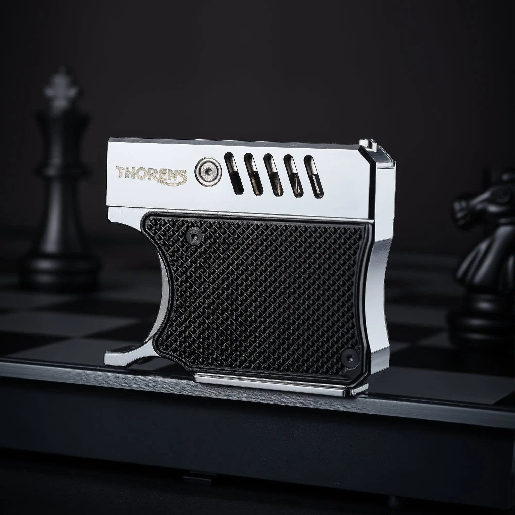 A side profile of the Thorens Gun Lighter standing on a chessboard. The image highlights the silver-polished metal upper frame, the textured black grip panel, and the unique vented barrel-style design.