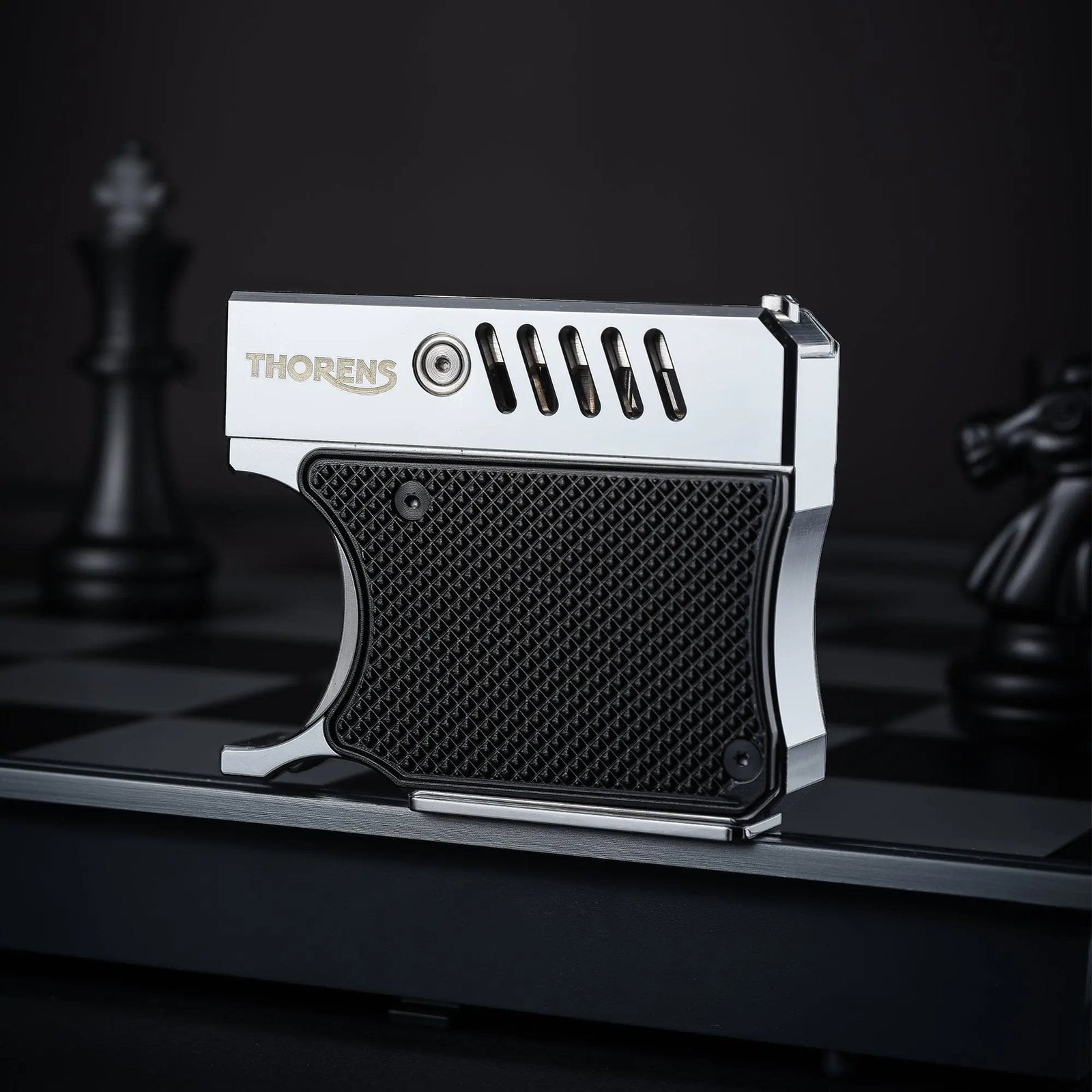A side profile of the Thorens Gun Lighter standing on a chessboard. The image highlights the silver-polished metal upper frame, the textured black grip panel, and the unique vented barrel-style design.
