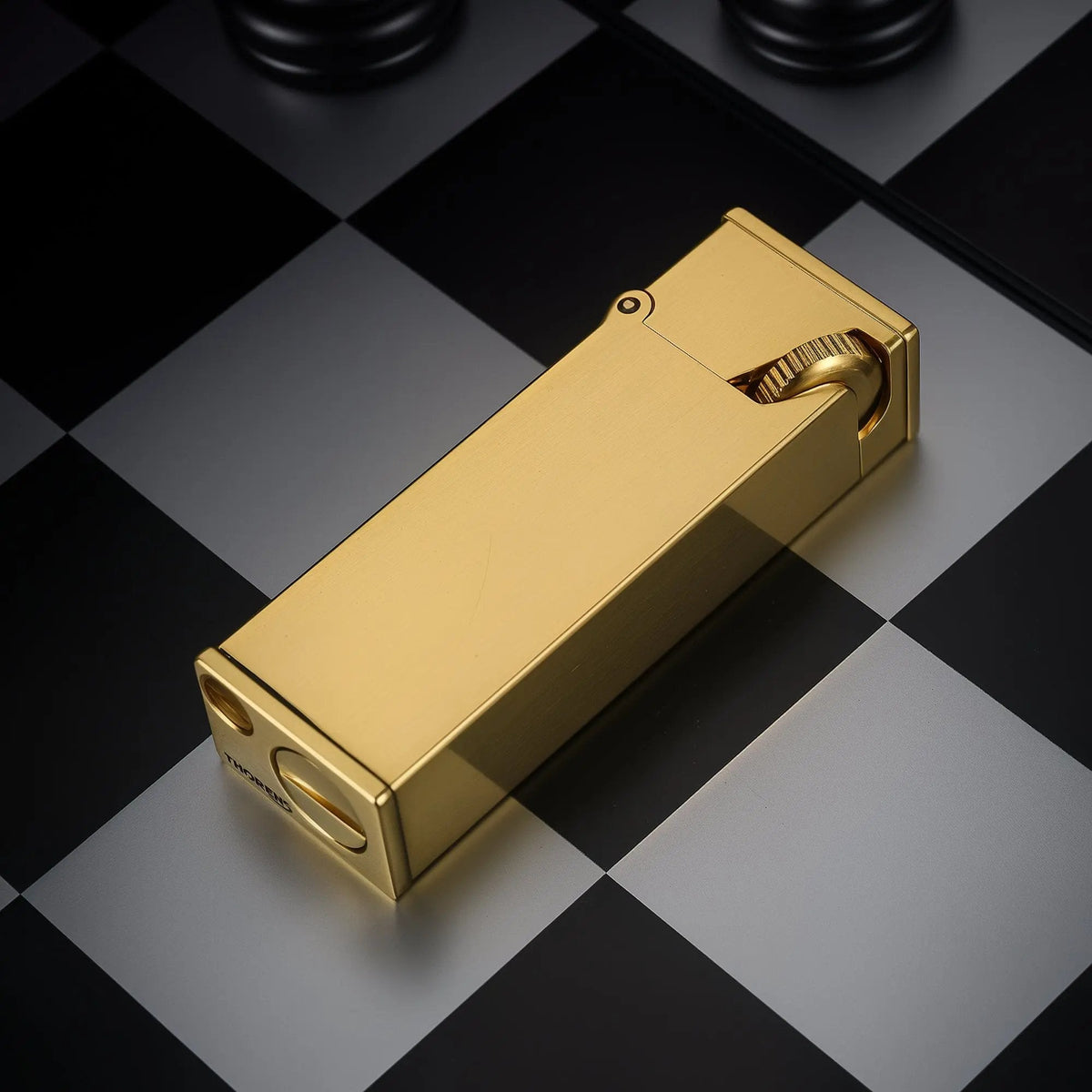 A glossy brass Long Fellow lighter positioned on a chessboard, showing the smooth rear finish, the base engraving, and the side profile of the inclined ignition wheel.
