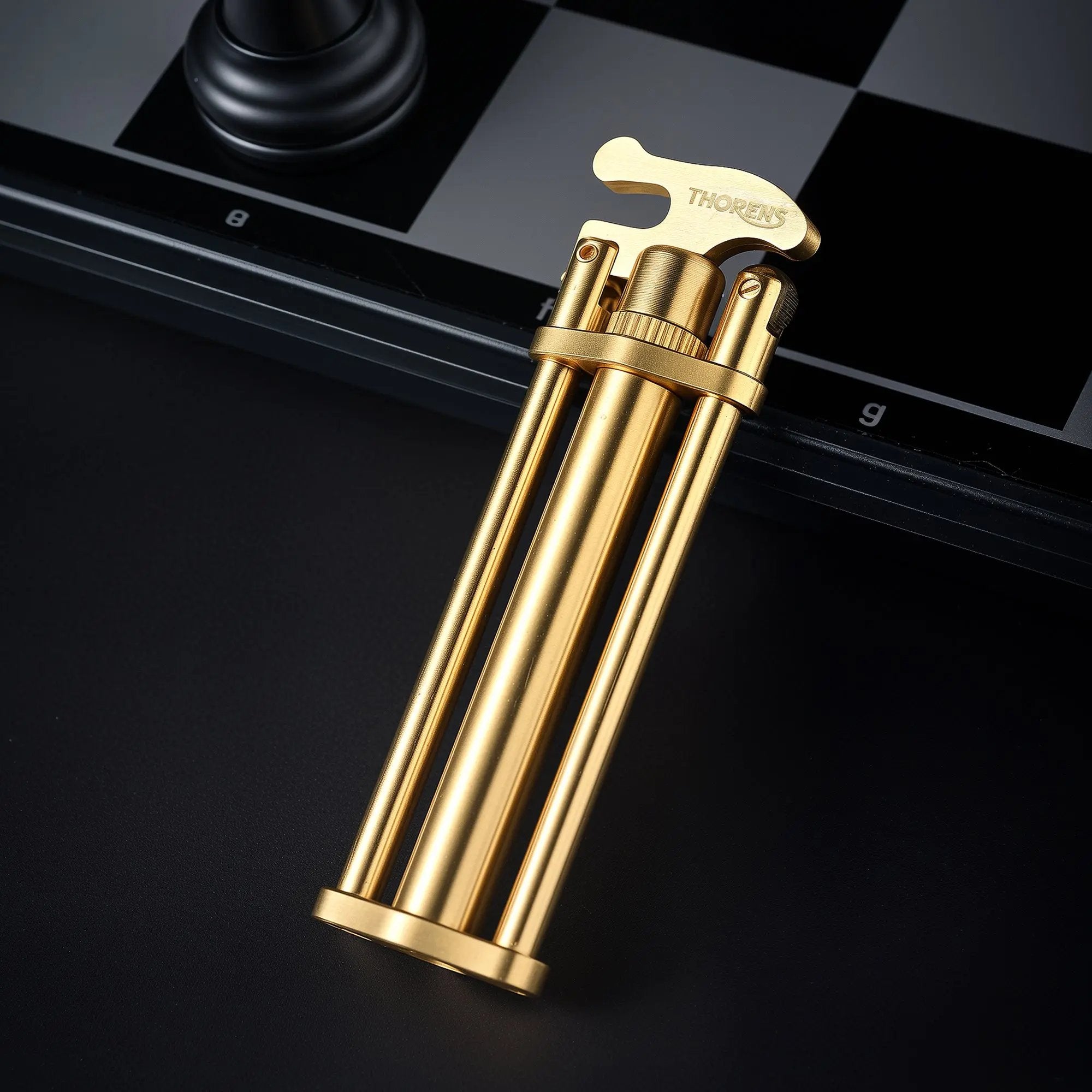 A standing Three-Section Stick Lighter featuring a brushed brass finish and engraved Thorens logo, positioned against a dark surface and chessboard.