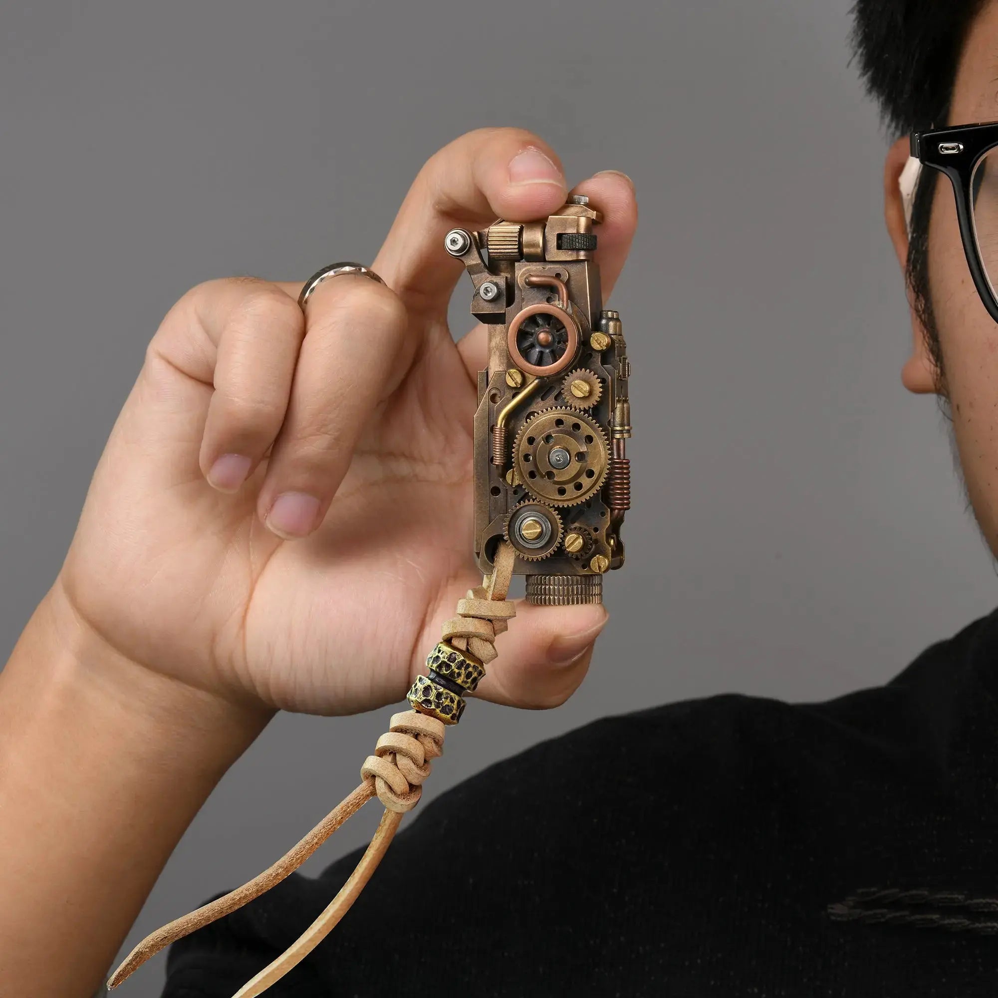 Thorens steampunk mechanical grip grand gear lighter in antique bronze held in hand, featuring oversized exposed gear detailing, mixed metal tones, and a leather lanyard with bead accents against a neutral background