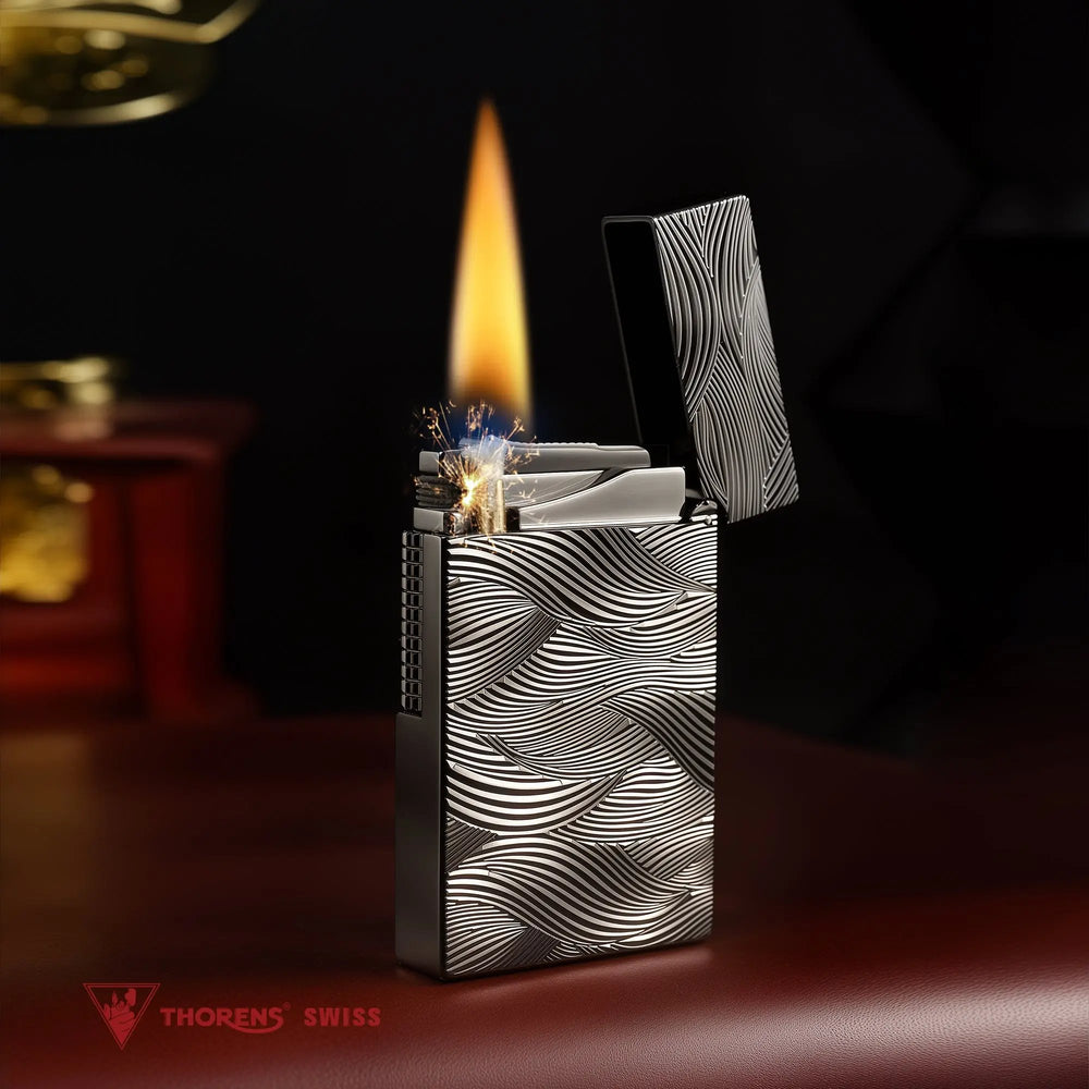 A black Aqua Rhythm lighter with an open lid is ignited, showing a steady flame and spark, featuring a dark brass body with silver wave engravings.