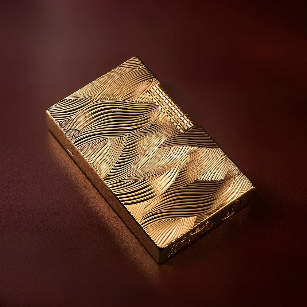 A gold Thorens Aqua Rhythm lighter resting on a rich mahogany surface. The design features a continuous, flowing wave pattern engraved across the entire face, with a prominent knurled side-roller.