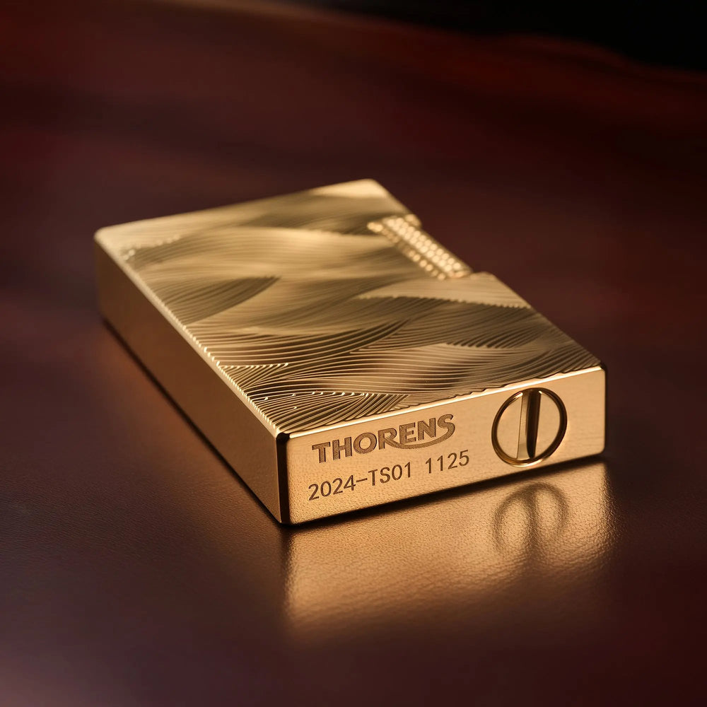 A close-up perspective of the gold Thorens Aqua Rhythm lighter lying on its side, highlighting the deep-etched wave patterns and the branded base featuring the Thorens logo, the "2024-TS01" serial series, and the fuel port.