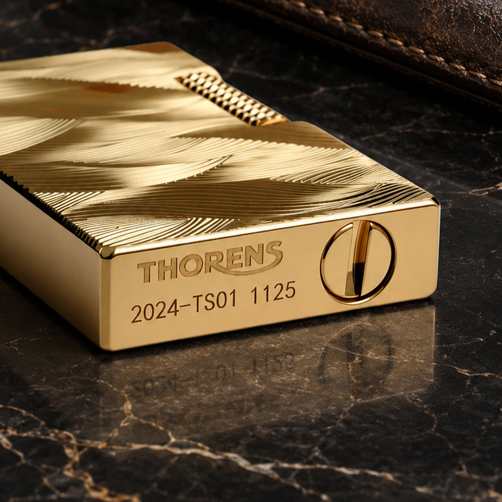 A close-up perspective of the gold Thorens Aqua Rhythm lighter lying on its side, highlighting the deep-etched wave patterns and the branded base featuring the Thorens logo, the "2024-TS01" serial series, and the fuel port.
