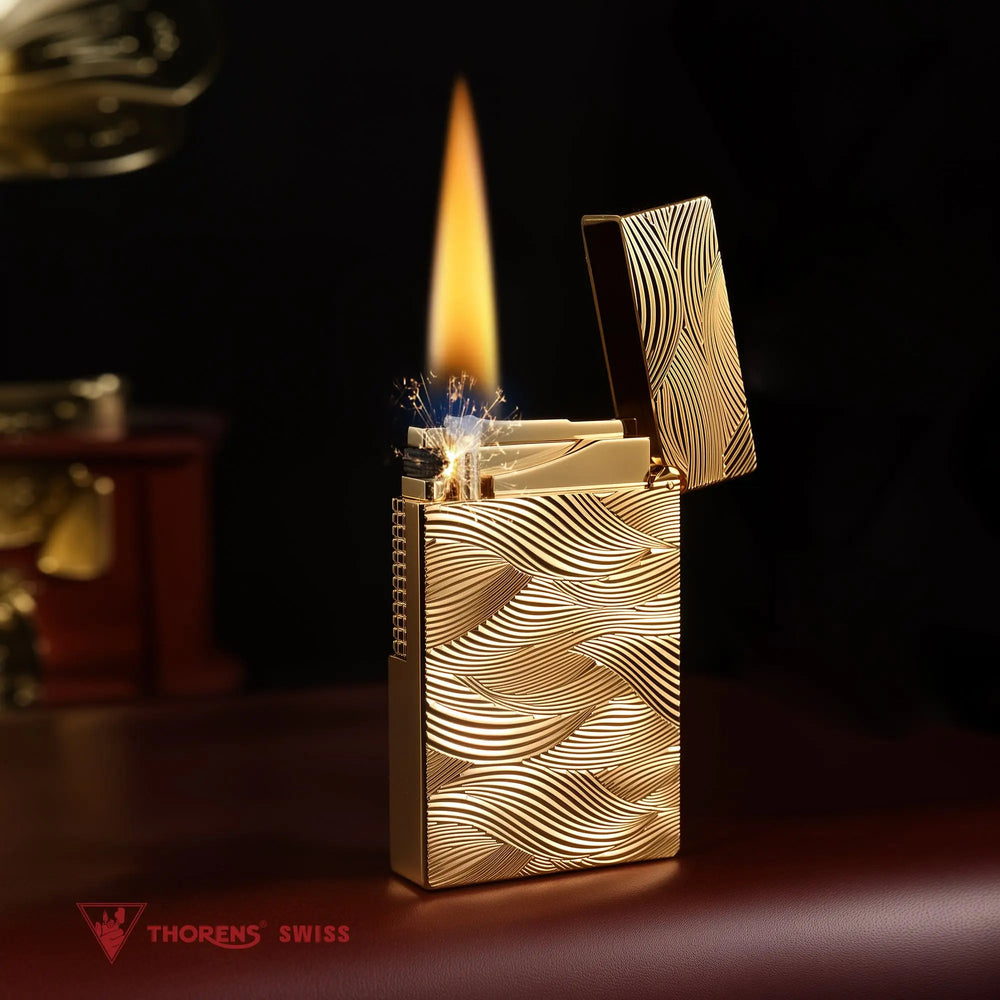An ignited gold Thorens Aqua Rhythm lighter showcasing a steady flame and active sparks. The deep wave engravings are beautifully illuminated by the fire, set against a dark, warm background.
