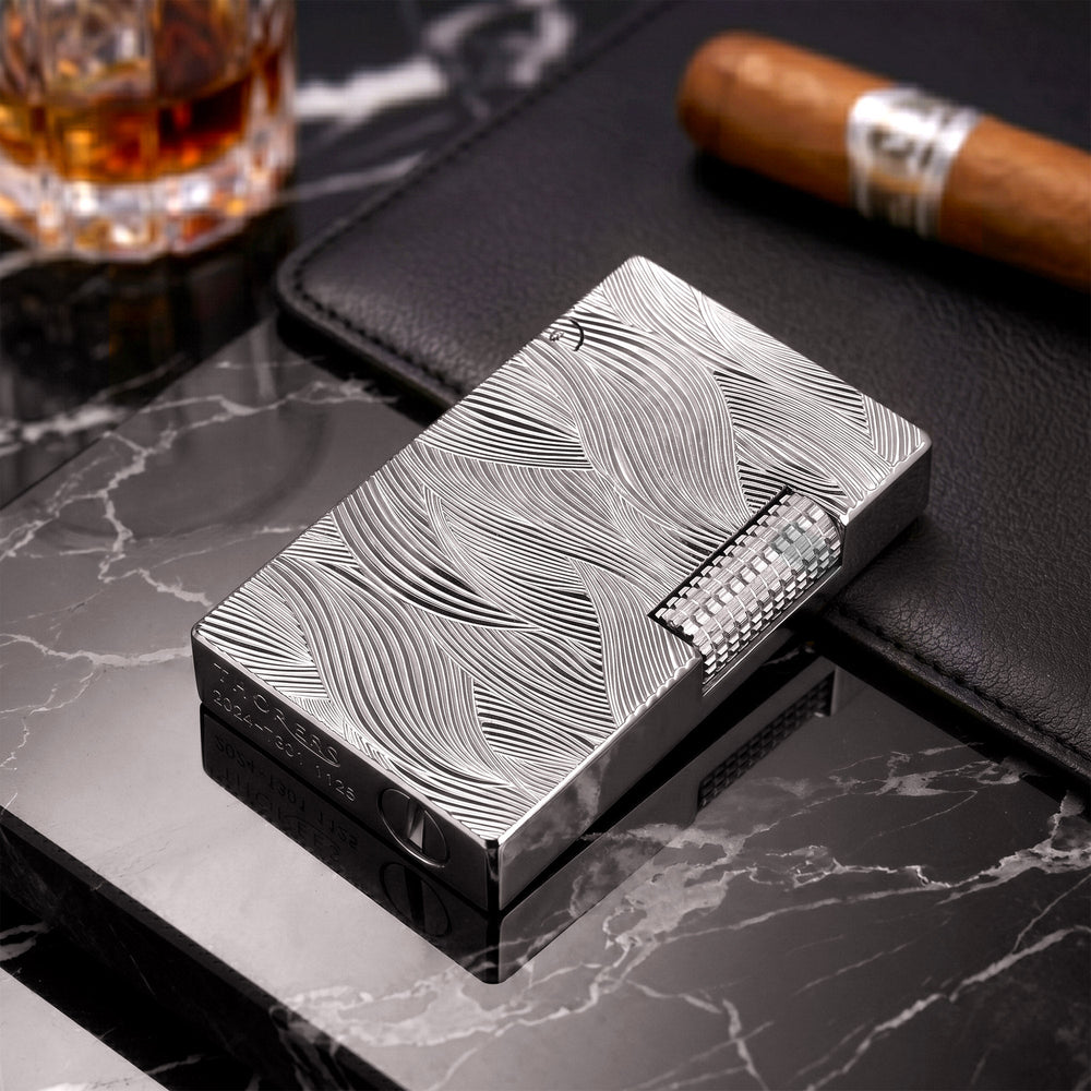 A silver Aqua Rhythm lighter with deep-etched wave patterns resting on a reflective surface, showcasing its metallic luster and precision craftsmanship.