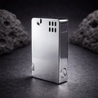 A high-angle studio photograph of the silver ASPREY-style lift-arm lighter standing on a dark, textured slate surface. The polished chrome-like finish reflects the light, highlighting its sleek, rectangular form and minimalist aesthetic. Visible features include the signature side-mounted striker wheel, the ventilation holes near the top, and the large flat-head screw at the base for fuel access. Two dark, volcanic-style rocks in the blurred background add a rugged, premium feel to the composition.