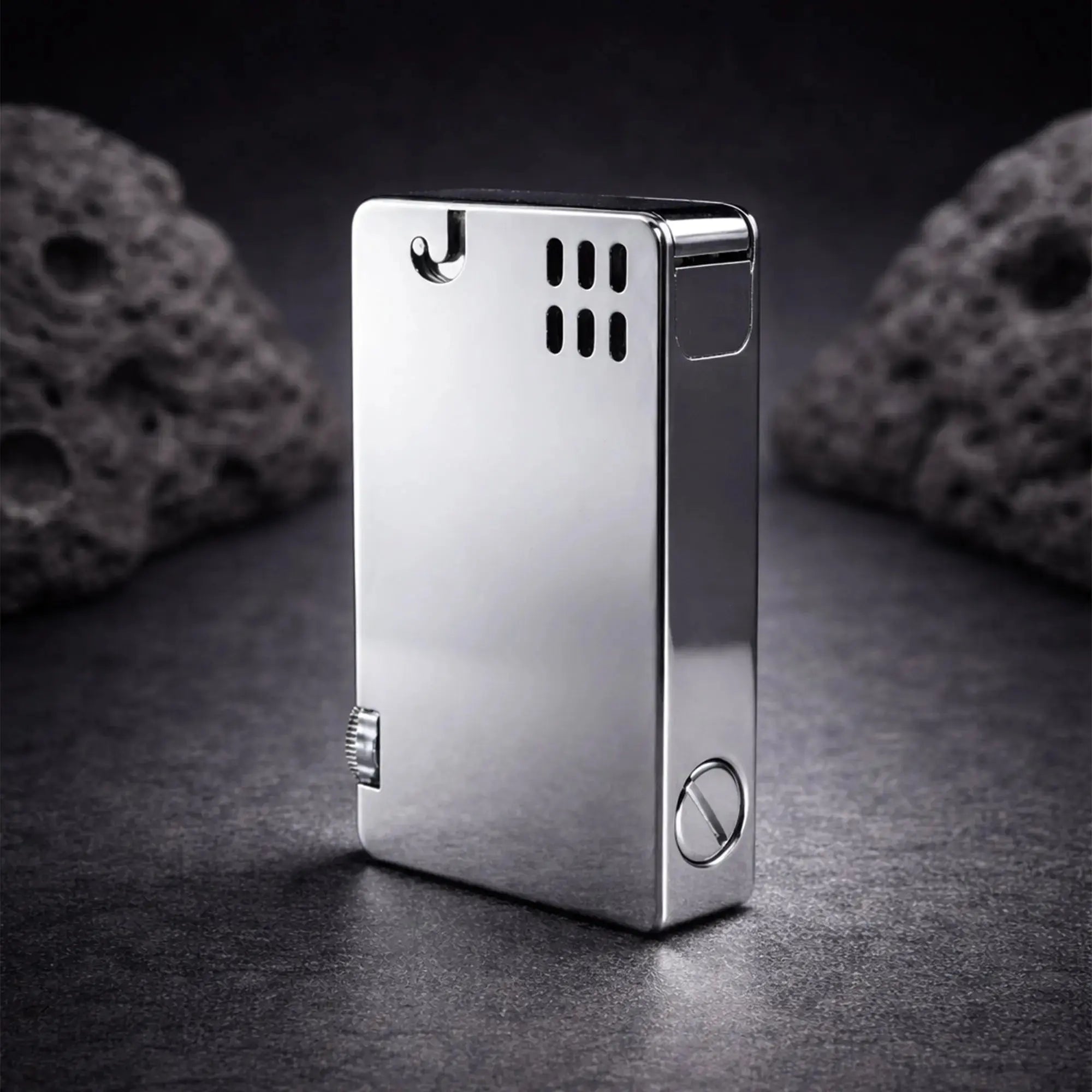 A high-angle studio photograph of the silver ASPREY-style lift-arm lighter standing on a dark, textured slate surface. The polished chrome-like finish reflects the light, highlighting its sleek, rectangular form and minimalist aesthetic. Visible features include the signature side-mounted striker wheel, the ventilation holes near the top, and the large flat-head screw at the base for fuel access. Two dark, volcanic-style rocks in the blurred background add a rugged, premium feel to the composition.