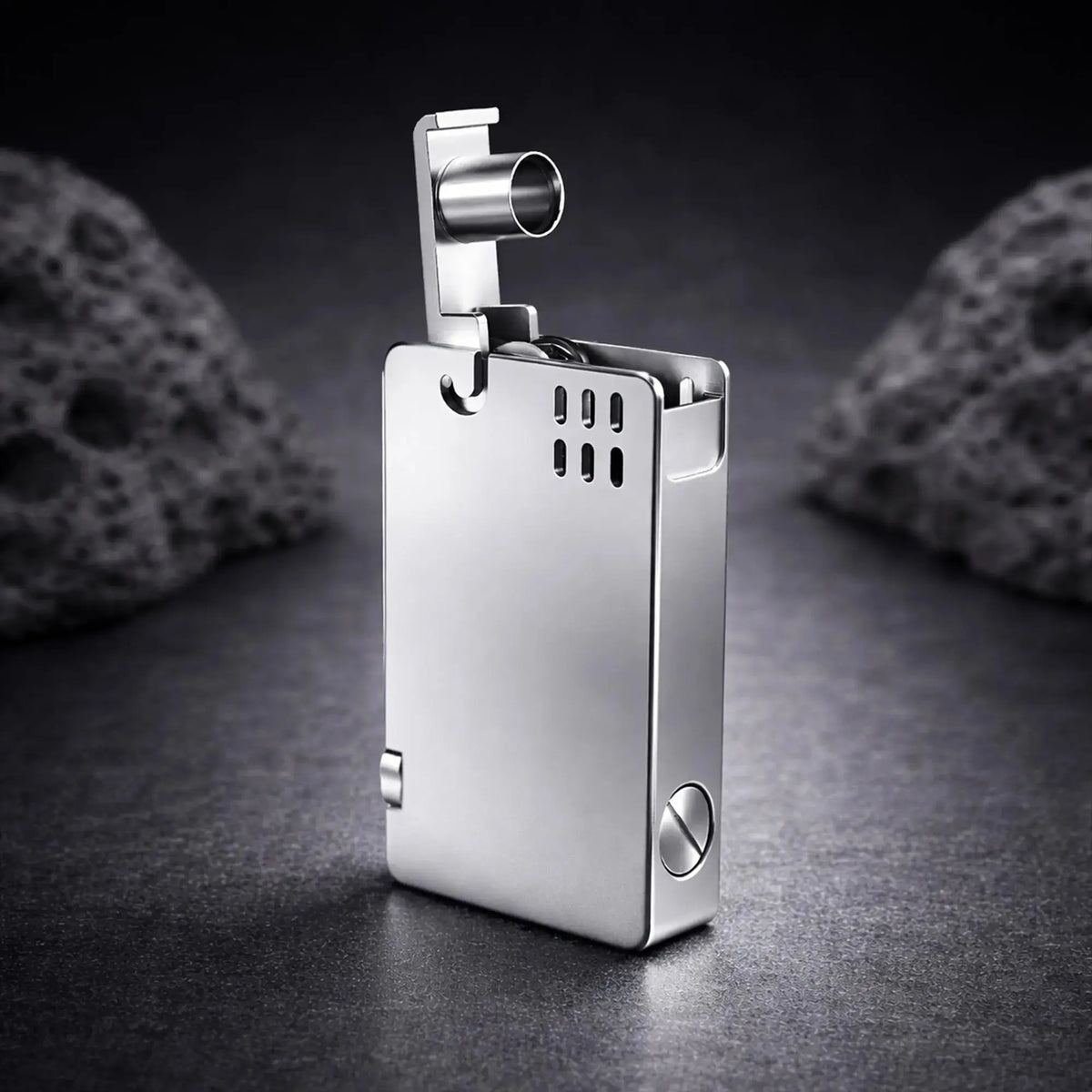 A high-angle studio photograph of the silver ASPREY-style lighter with the lift-arm mechanism fully raised. The open arm reveals the cylindrical snuff cap and exposes the internal ignition area where the wick and striker wheel are located. The polished chrome-like finish reflects the light, showcasing the sleek, rectangular body and minimalist ventilation holes against a dark, textured slate background with blurred volcanic rocks, emphasizing the lighter's precision mechanical design.