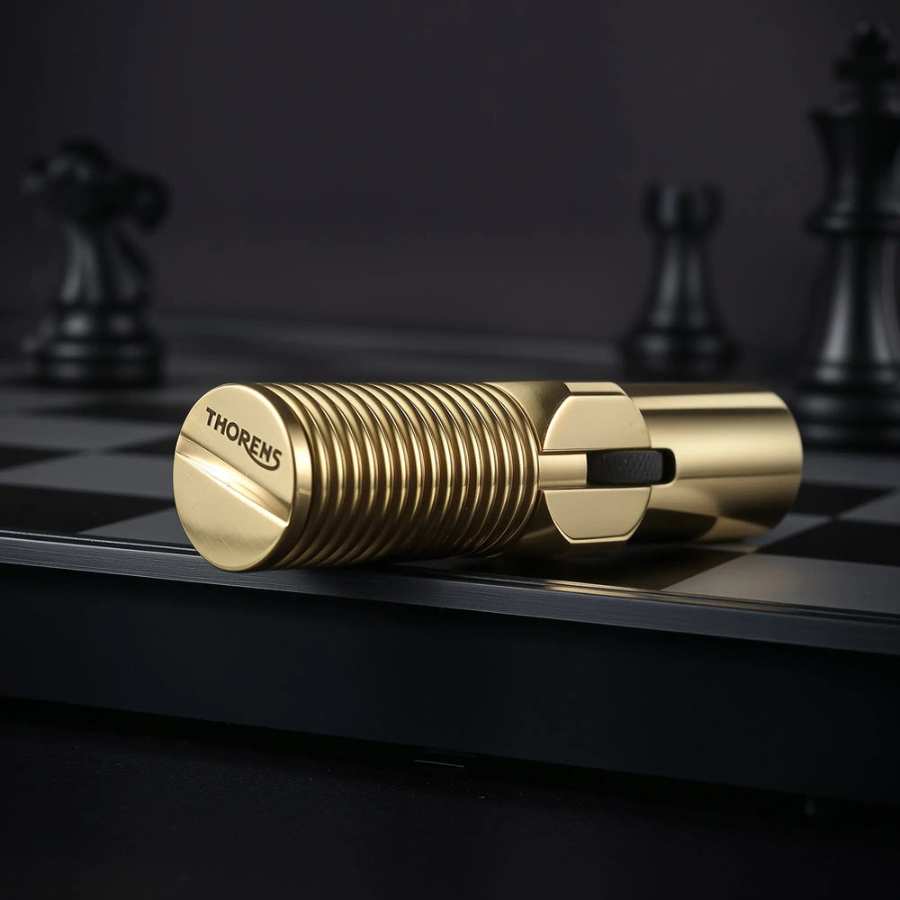 A horizontal close-up of the brass Big Screw Trench Lighter on a black chessboard, prominently displaying the "THORENS" logo engraved on the flat screw-head base.