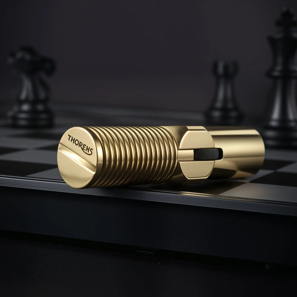 A horizontal close-up of the brass Big Screw Trench Lighter on a black chessboard, prominently displaying the "THORENS" logo engraved on the flat screw-head base.