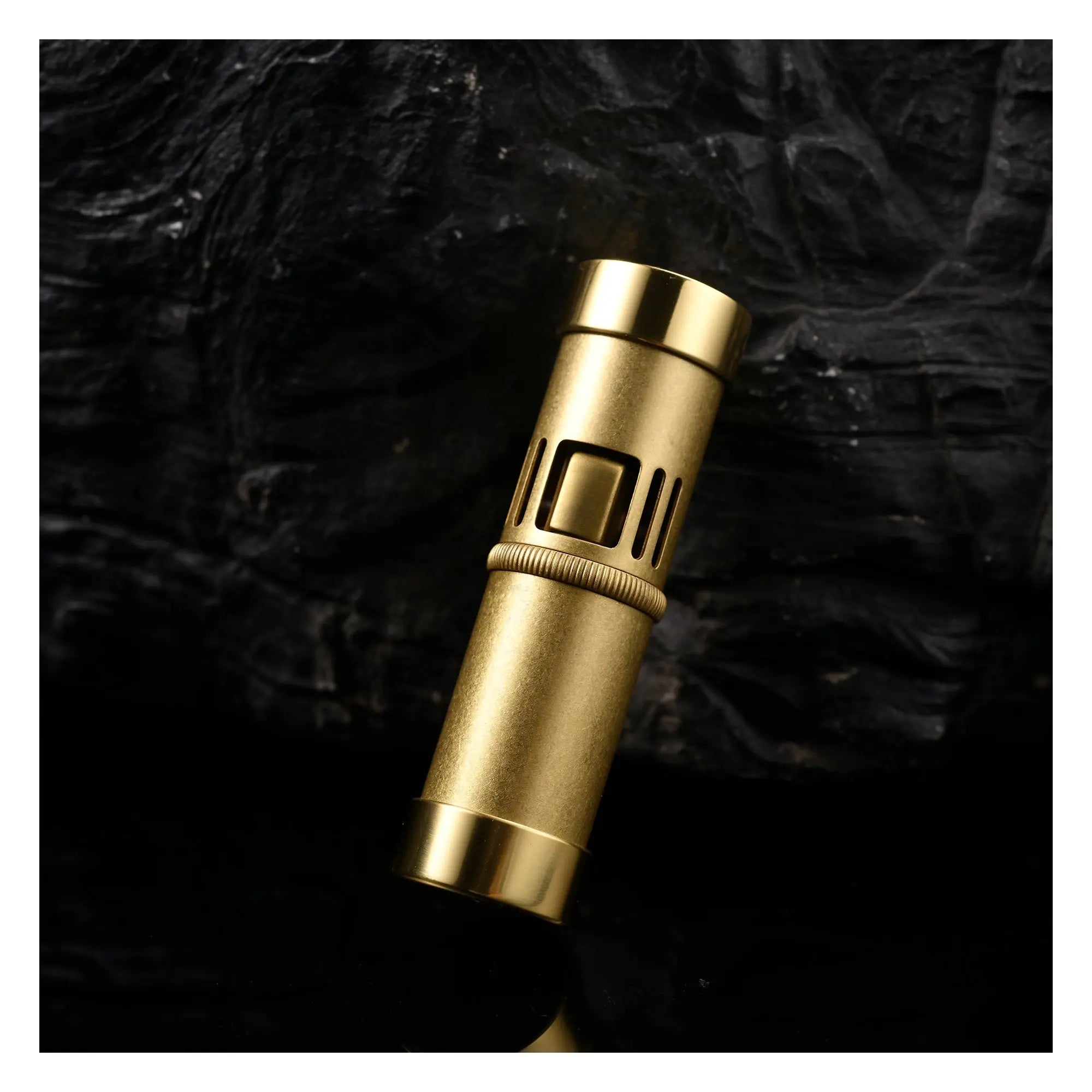 A Thorens Blaze Capsule lighter in vintage brass, standing against a dark, rugged stone background that emphasizes its durable construction and industrial aesthetic.