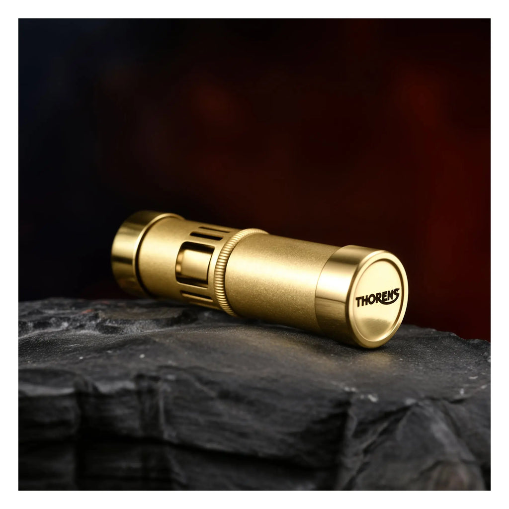 A horizontal view of the Thorens Blaze Capsule lighter in vintage brass, resting on a dark rugged rock surface, showcasing the "THORENS" logo engraved on the base cap.