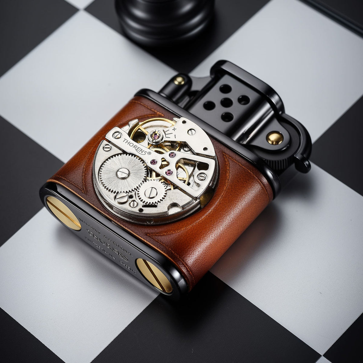 Thorens handmade cowhide lighter in brown and black resting on a chessboard surface, showing inlaid mechanical movement, leather body, and brass base screws.