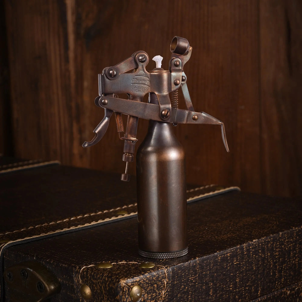 A Capitol Table Lighter with a glossy bronze finish and complex lever mechanism, standing on a leather-textured trunk against a rustic wooden background.