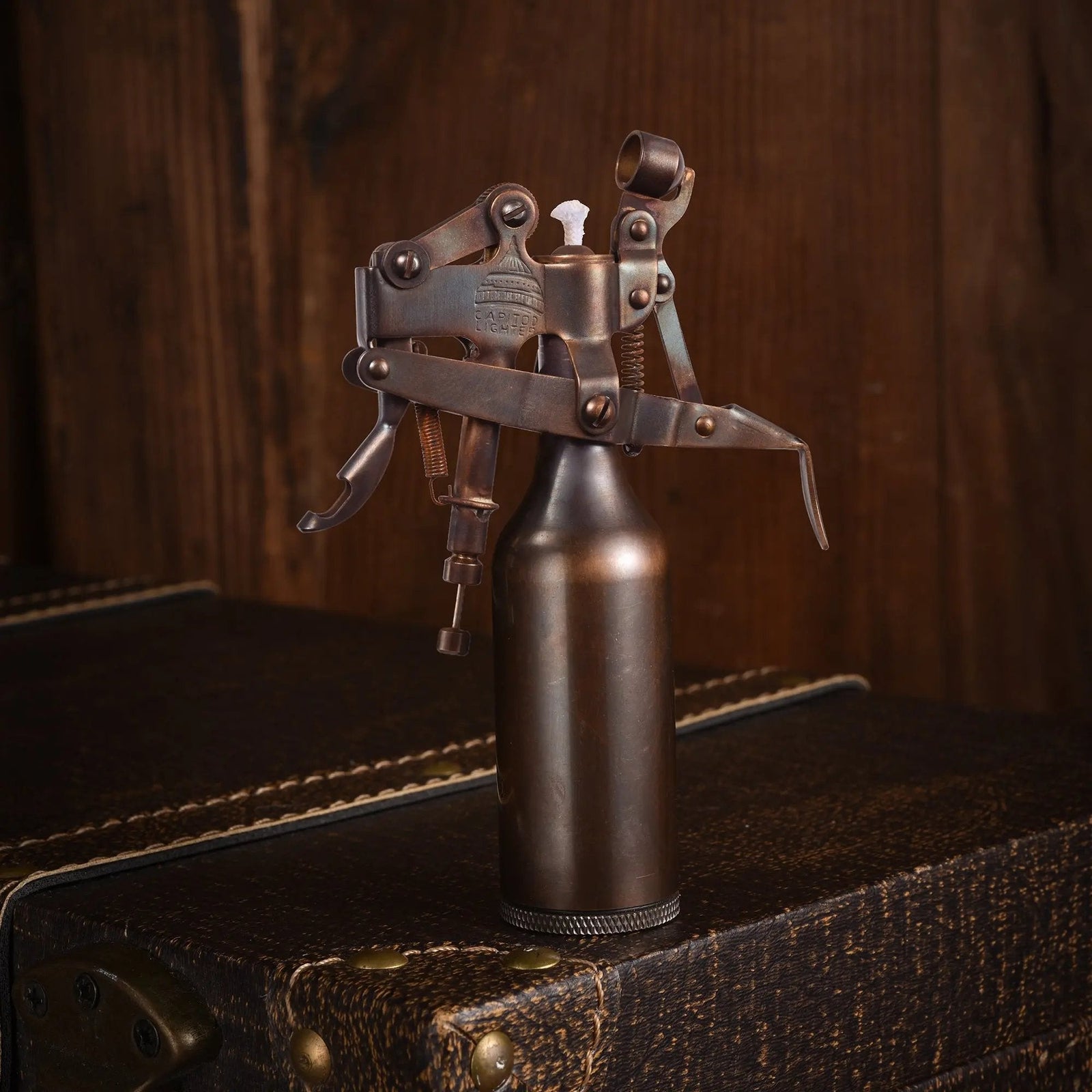A Capitol Table Lighter with a glossy bronze finish and complex lever mechanism, standing on a leather-textured trunk against a rustic wooden background.