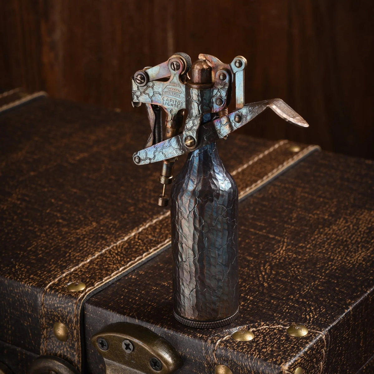 A hand-carved Capitol Table Lighter with a distressed, crackled texture and iridescent antique patina, standing on a dark leather vintage trunk against a wooden backdrop.