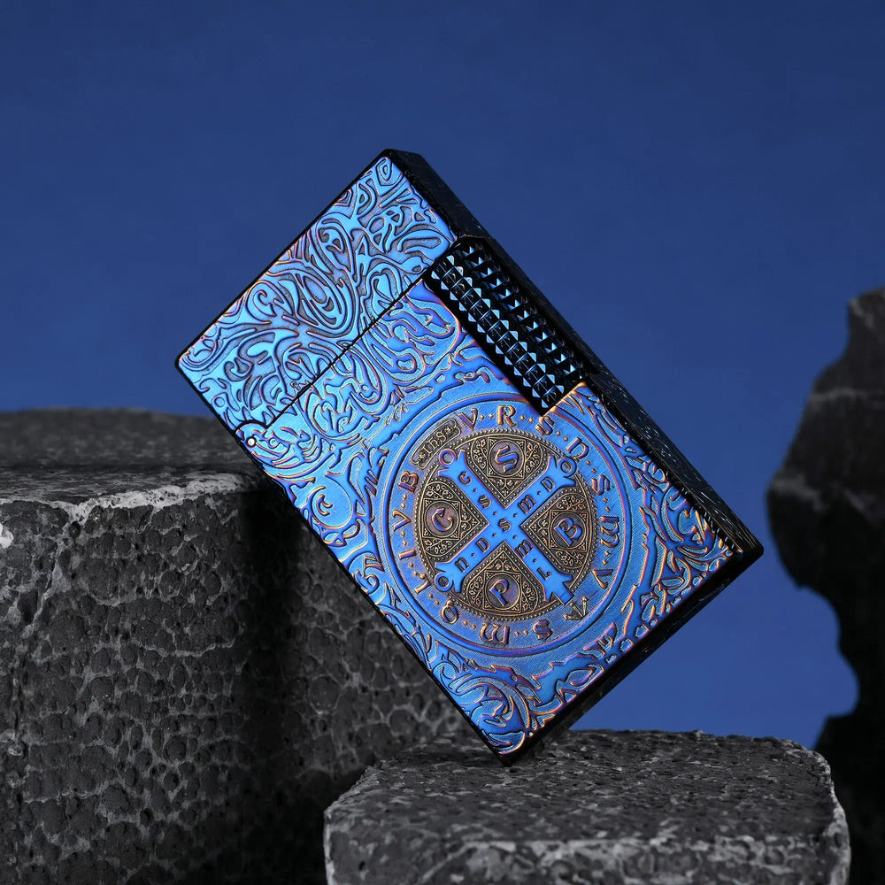 A closed Constantine Midnight Blue Sigil lighter leaning against volcanic stone, showcasing its deep-etched scrollwork, textured side roller, and central circular Saint Benedict sigil in a vibrant blue and gold-tone finish.