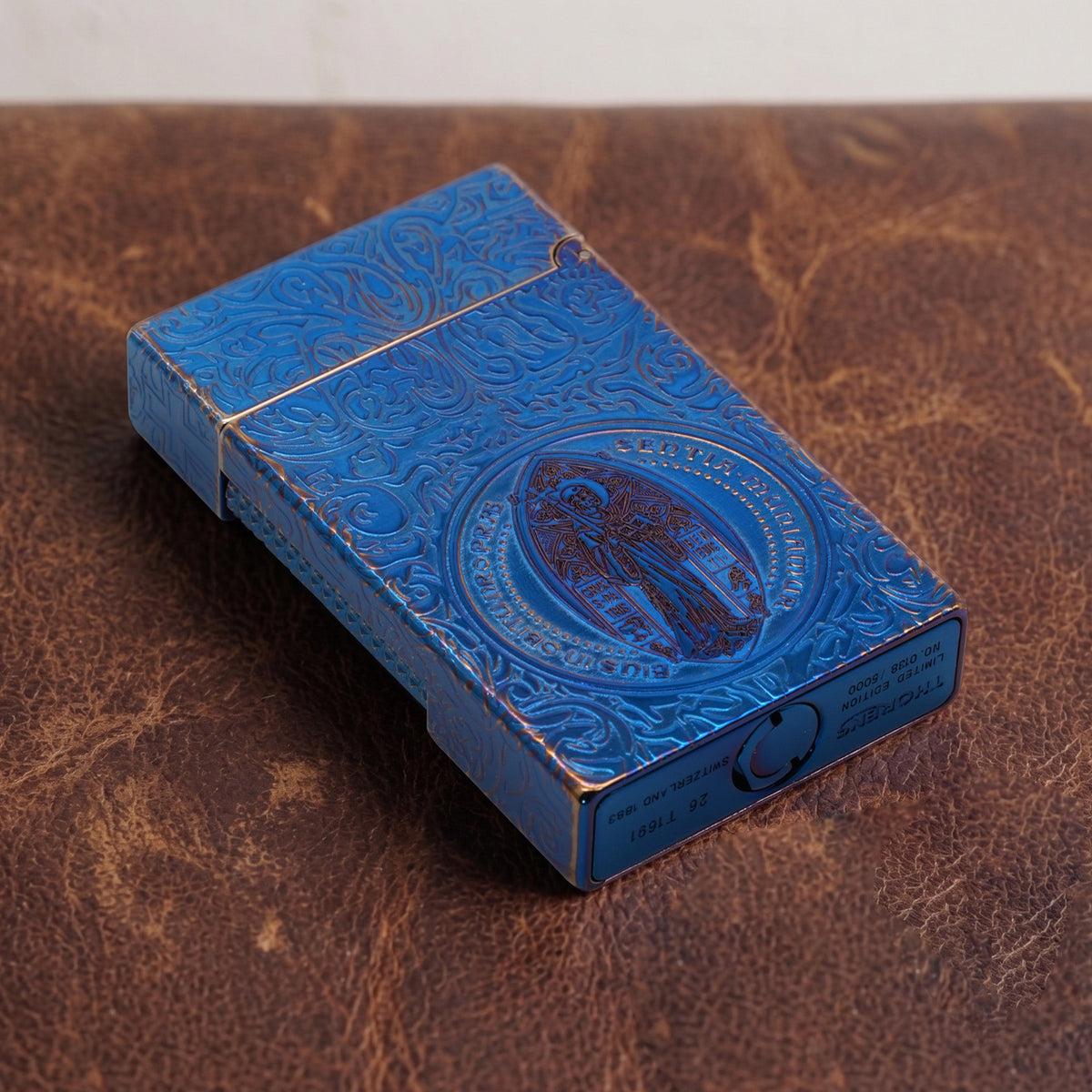 A closed Constantine Midnight Blue Sigil lighter leaning against volcanic stone, showcasing its deep-etched scrollwork, textured side roller, and central circular Saint Benedict sigil in a vibrant blue and gold-tone finish.