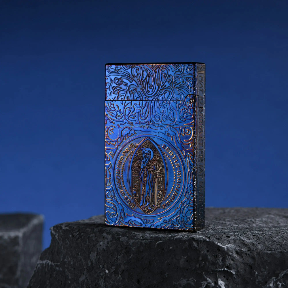 A closed Constantine Midnight Blue Sigil lighter featuring deep-etched gothic engravings and a central religious sigil, resting on a dark, textured volcanic rock against a blue gradient background.