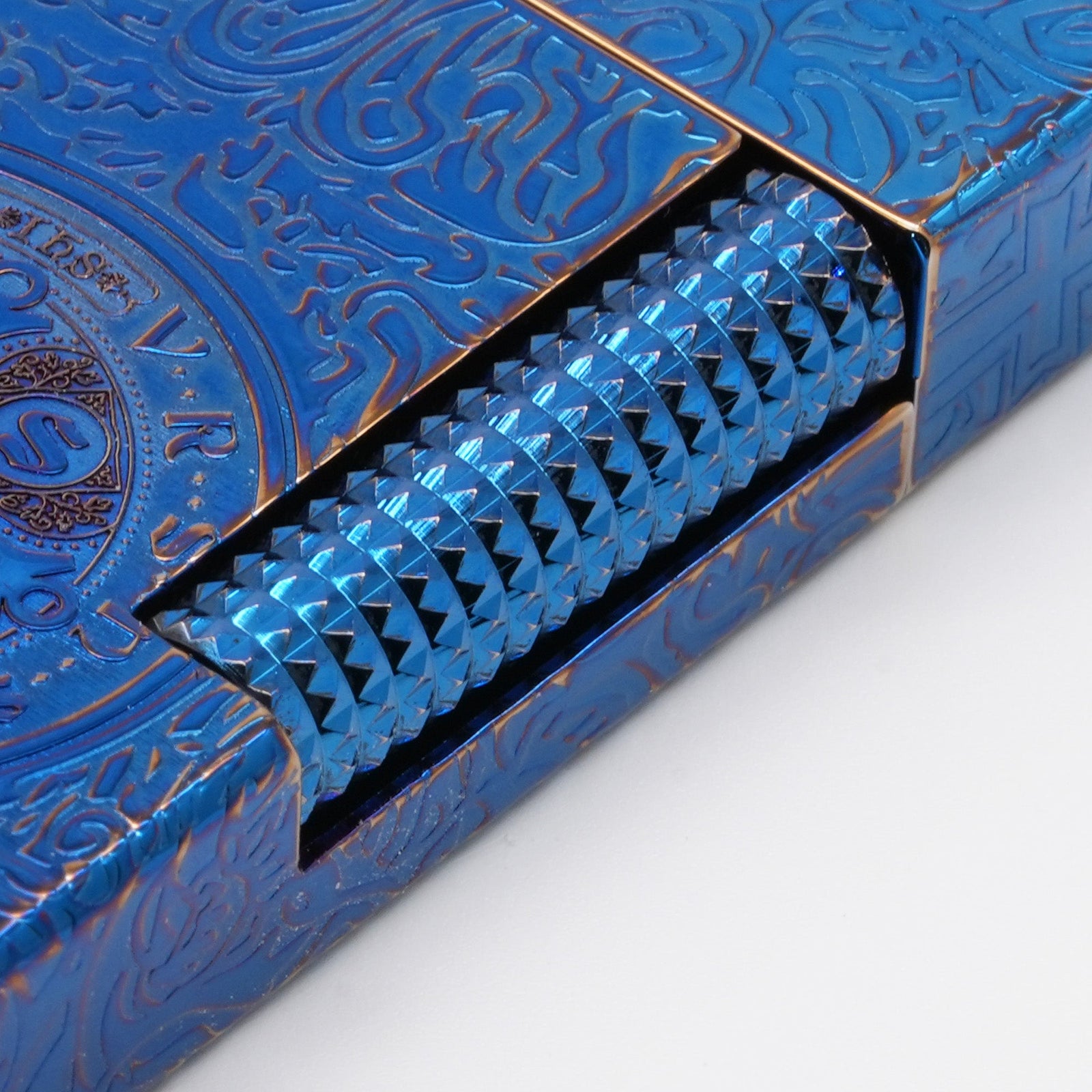 A closed Constantine Midnight Blue Sigil lighter featuring deep-etched gothic engravings and a central religious sigil, resting on a dark, textured volcanic rock against a blue gradient background.