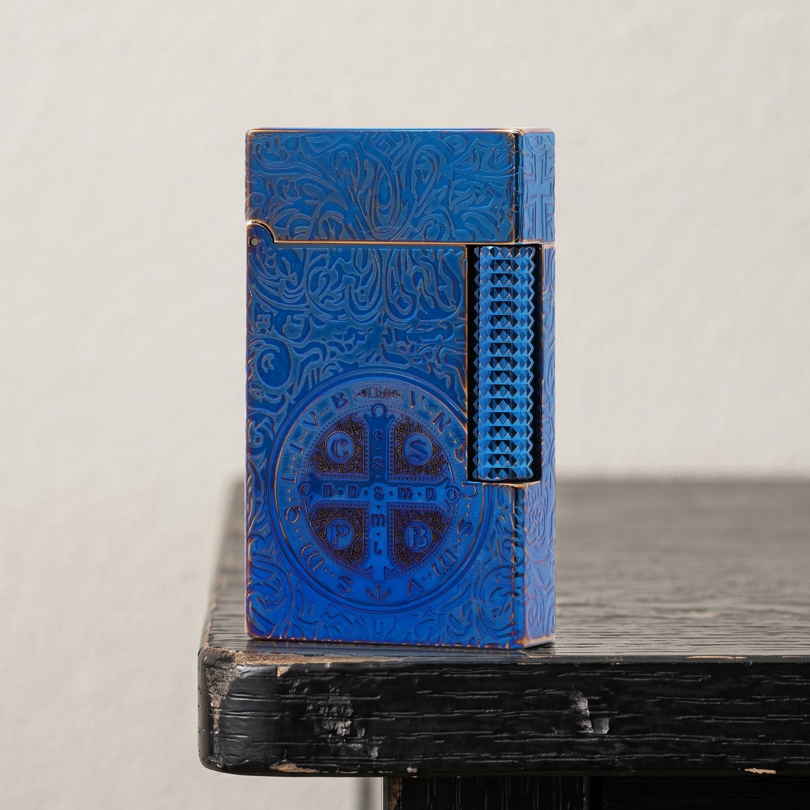 An open, ignited Constantine Midnight Blue Sigil lighter stands vertically, showcasing its detailed gothic engravings and prominent religious sigil against a dark, blue-lit background with a large cocktail glass.