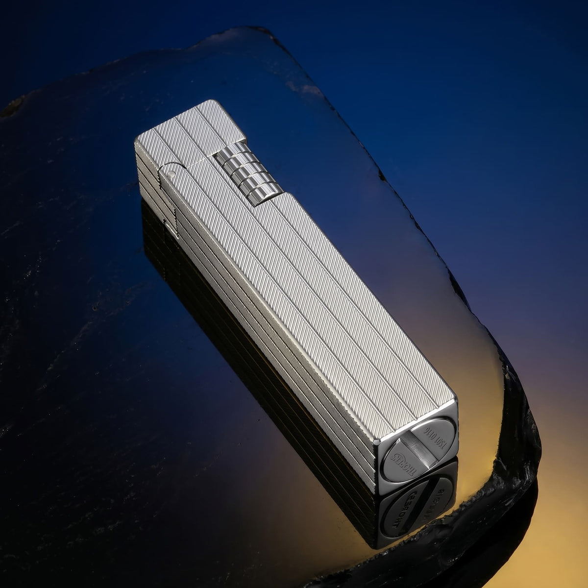 Thorens rectangular lighter in silver placed at an angle, featuring diagonal engraved brass surface, elongated form, and side ignition wheel on a dark reflective surface.