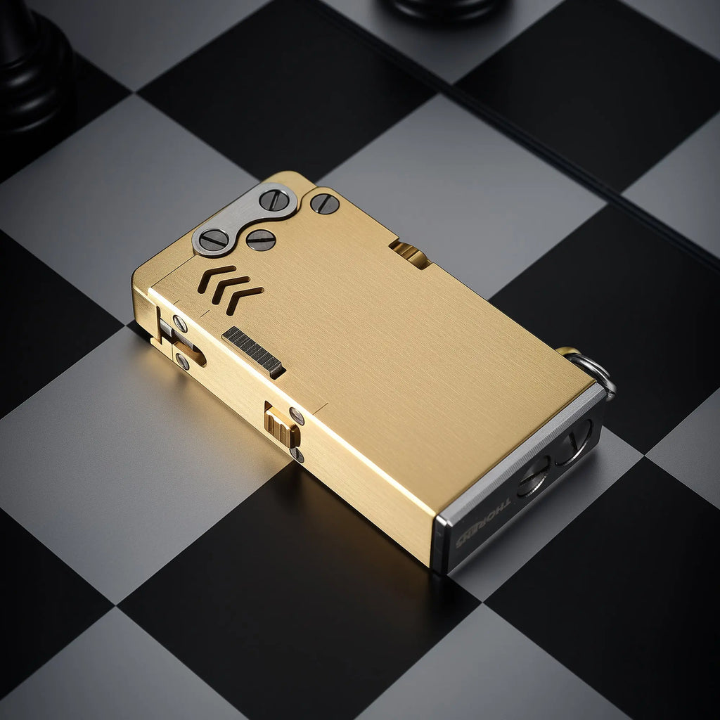 Thorens Ejection Lighter in brushed brass finish resting closed on a chessboard surface, showing engraved vents and precision mechanical construction.