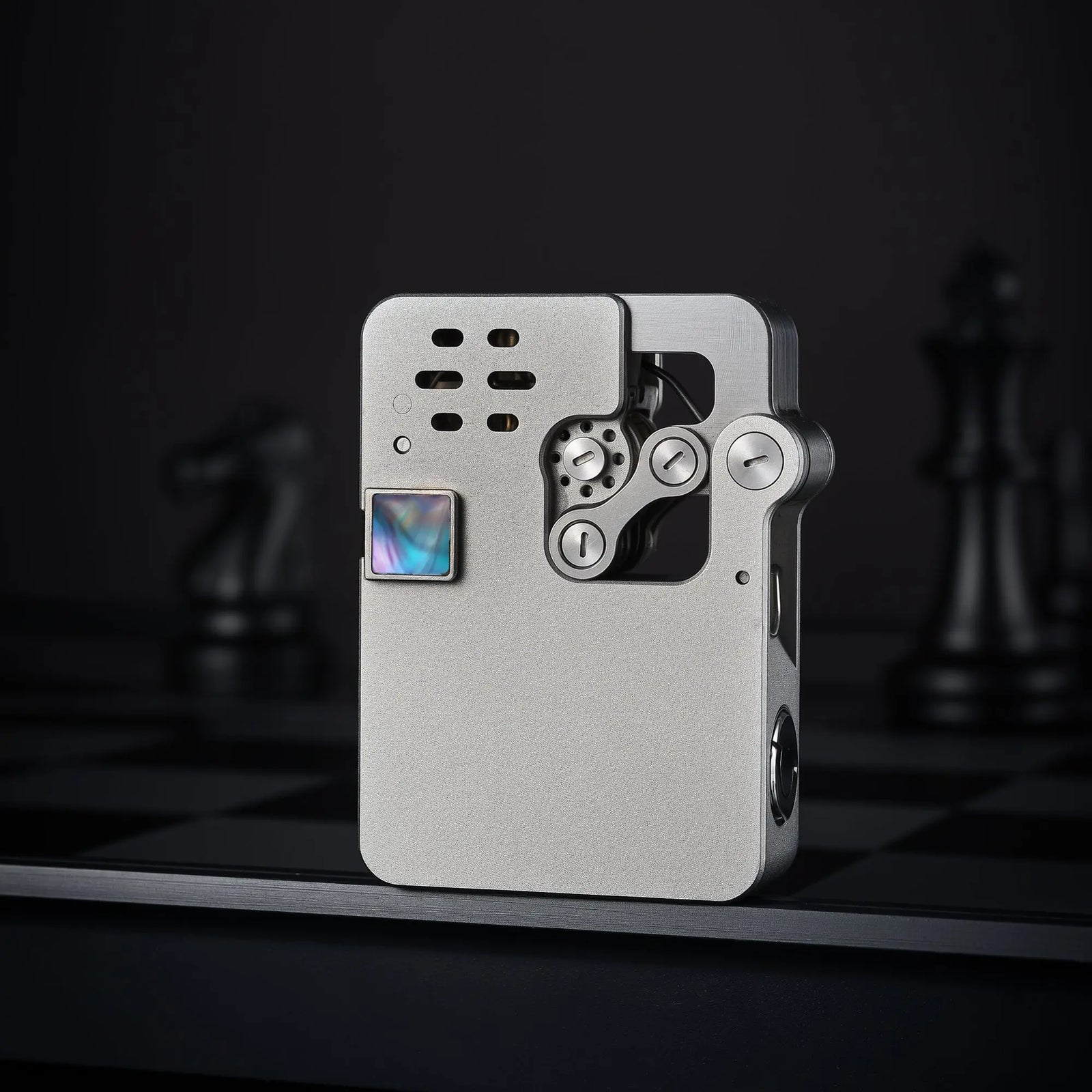 A front-view of the matte grey Ejection Lighter Hollowing Machine, displaying its mechanical gears, linkage bars, and square iridescent inlay on a dark chessboard.